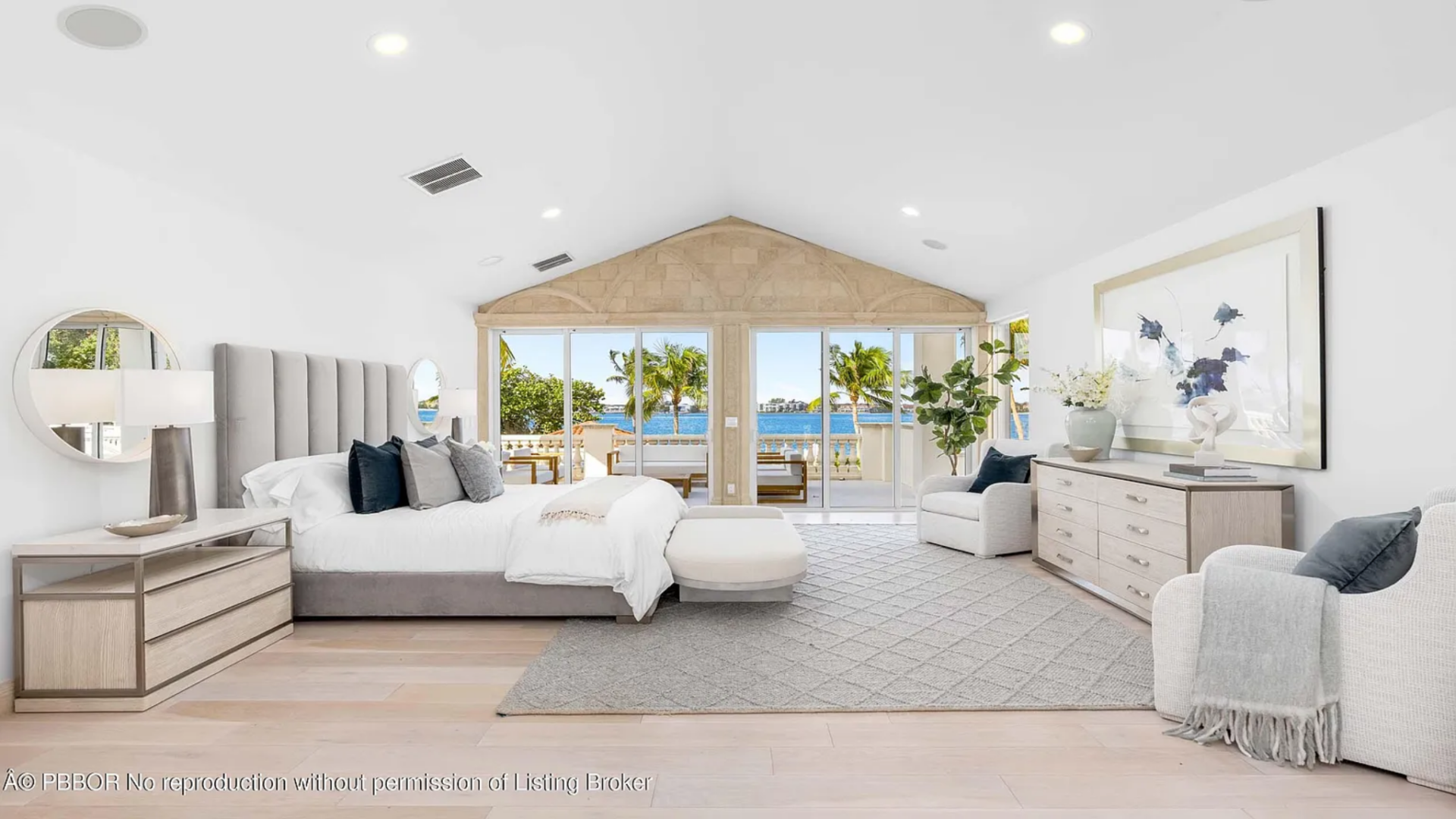 A large upholstered bed with a tall, vertically paneled headboard anchors the space, framed by matching nightstands, table lamps, and round mirrors mounted on the wall above.