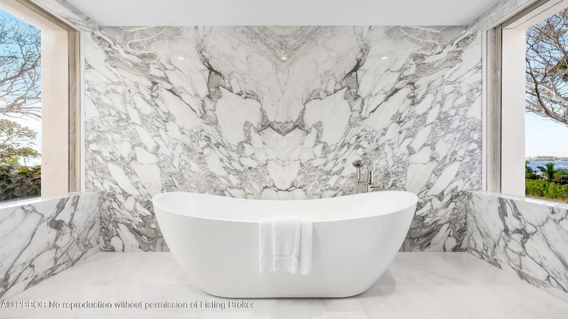 At the far end of the space, a freestanding soaking tub sits in front of a dramatic bookmatched marble wall where the stone’s pattern mirrors across the center, creating a striking focal point.