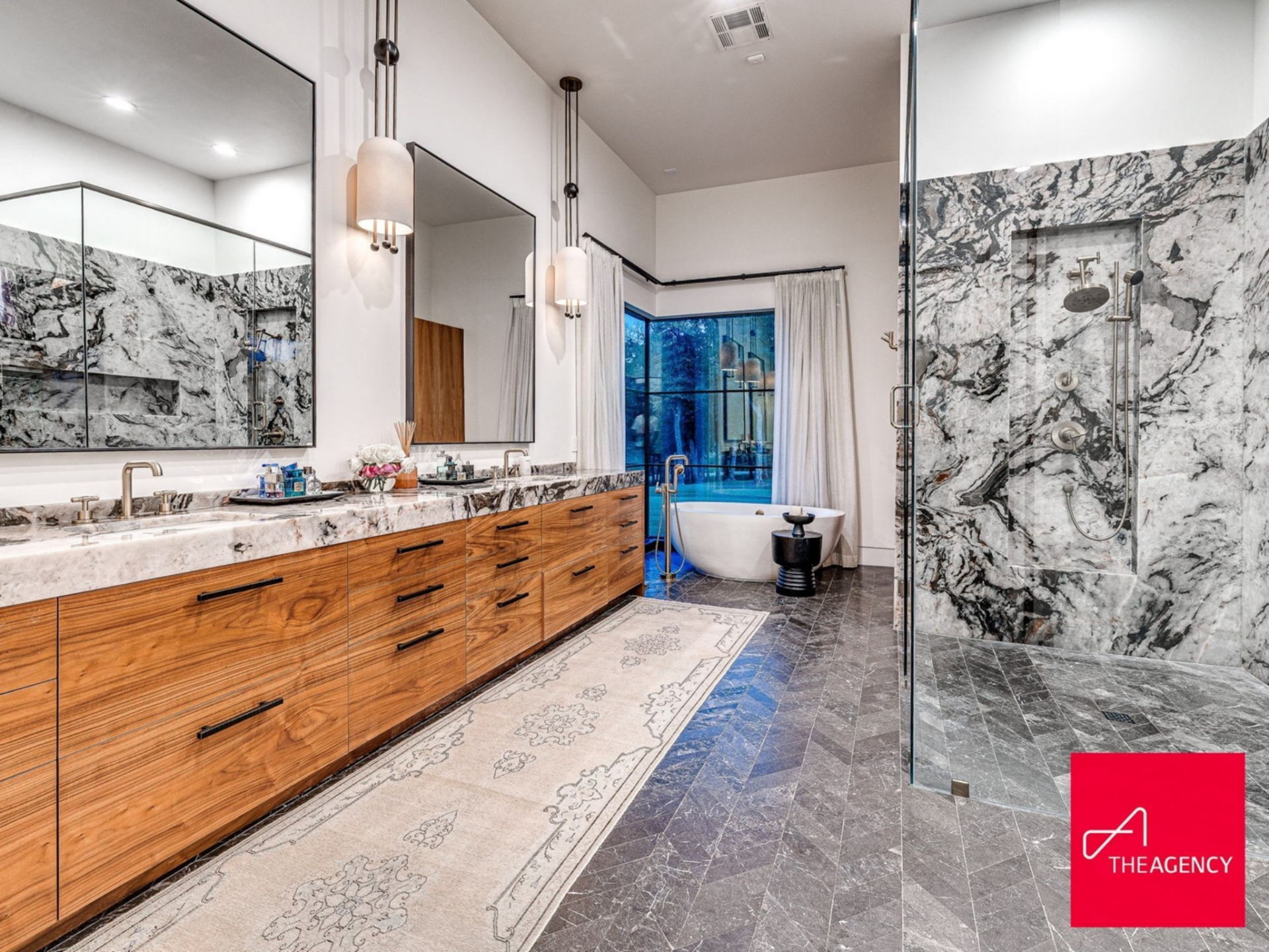 Across the room, a freestanding soaking tub sits in front of a window, positioned to capture outdoor views, while a glass-enclosed shower is wrapped in striking slab stone.