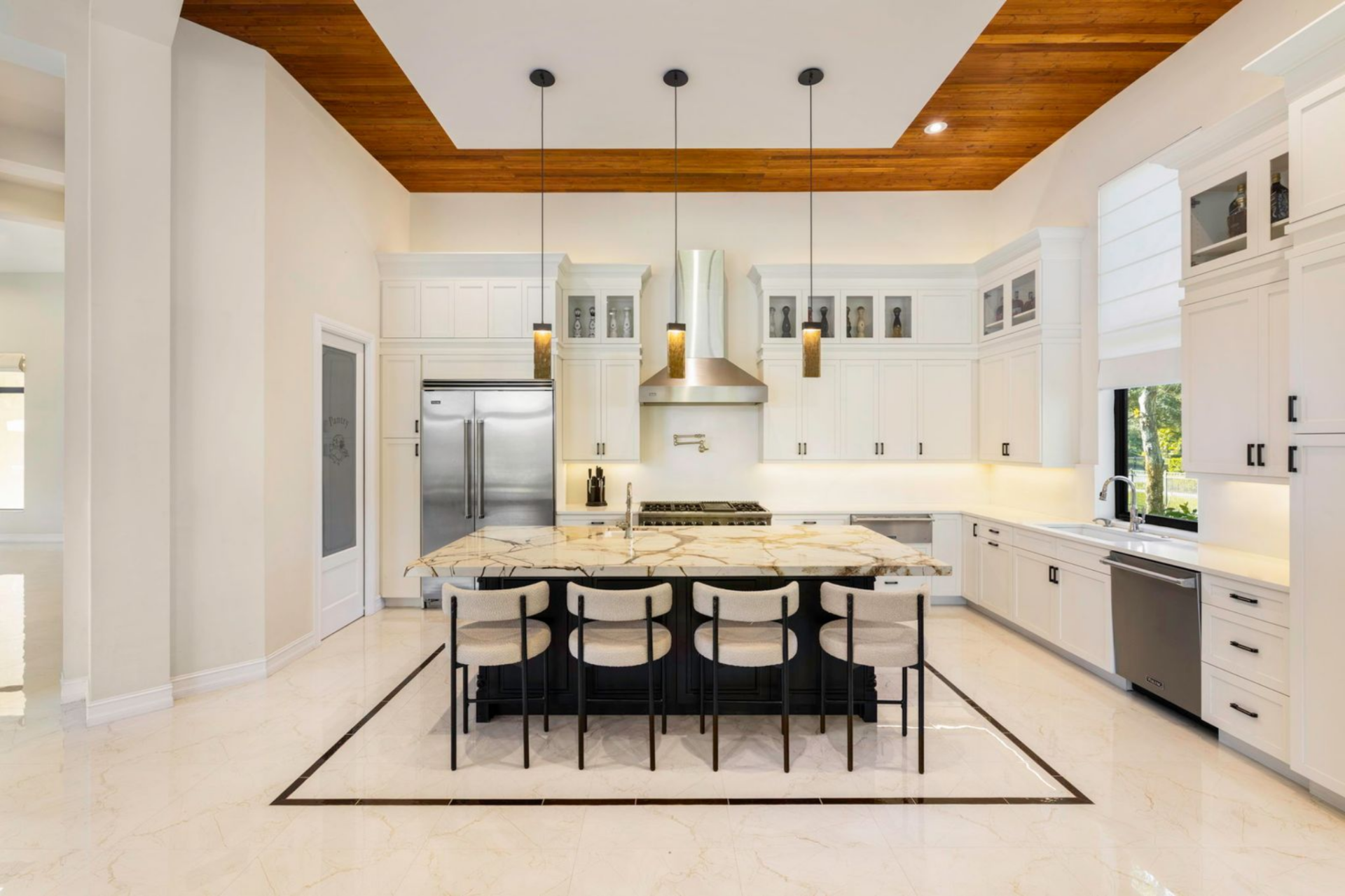 At the center sits an oversized marble island with seating for four, while white cabinetry stretches across the walls beneath a tray ceiling finished with warm wood inlays.