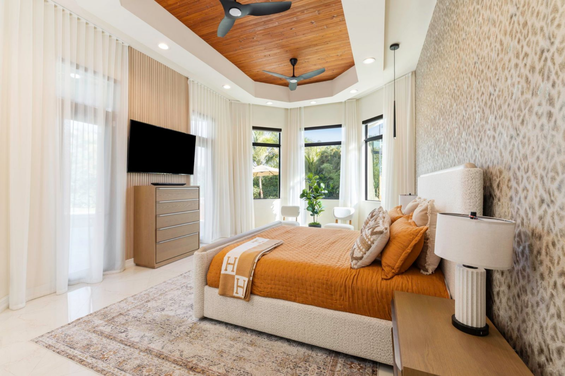 A tray ceiling finished with warm wood paneling and dual ceiling fans adds architectural detail above the bed, while a textured accent wall and neutral furnishings keep the space calm and refined.