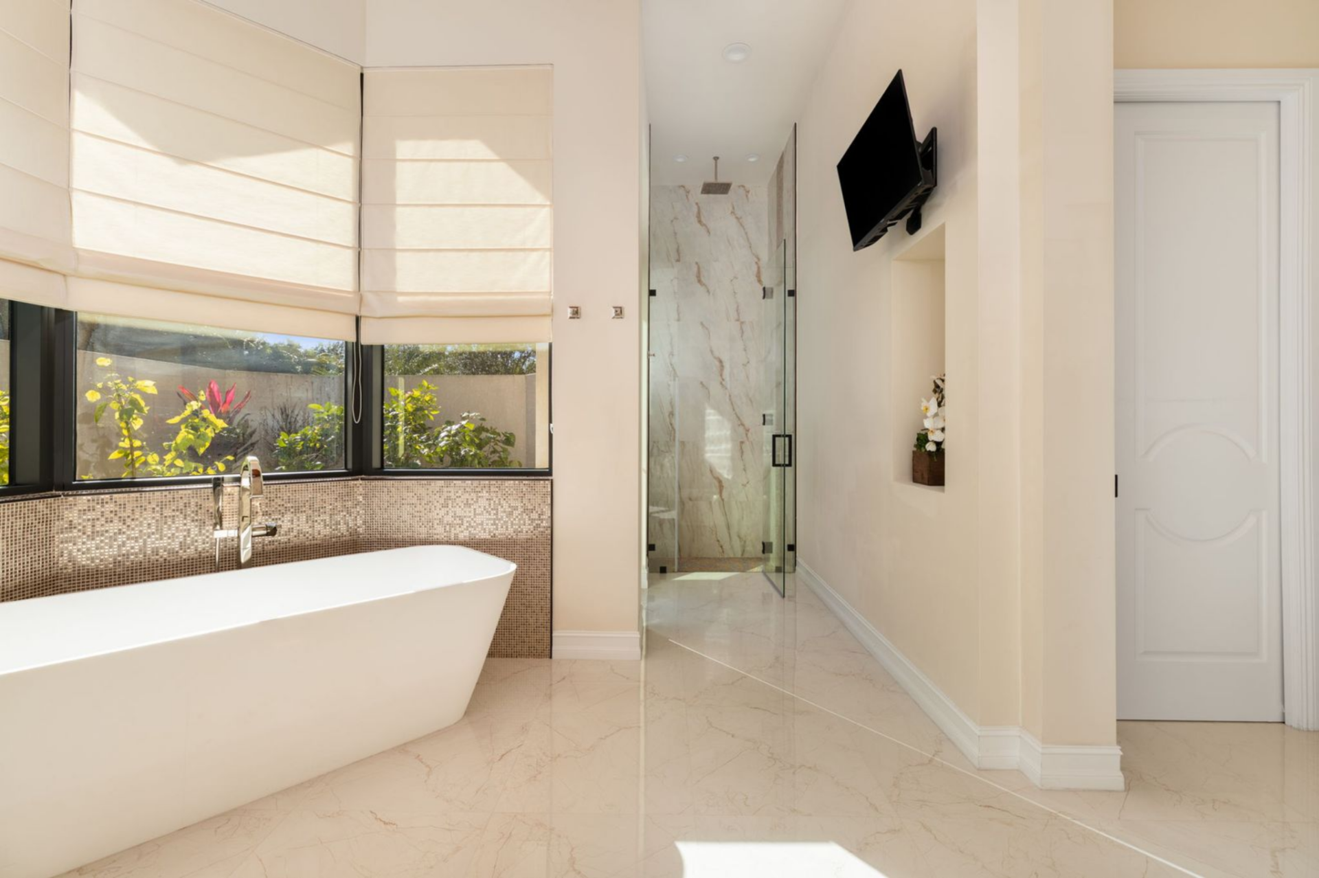 The spa-inspired bathroom continues the same bright, open feel with polished floors, a freestanding soaking tub positioned beneath a wide window, and a large glass-enclosed shower finished with marble-style walls. 