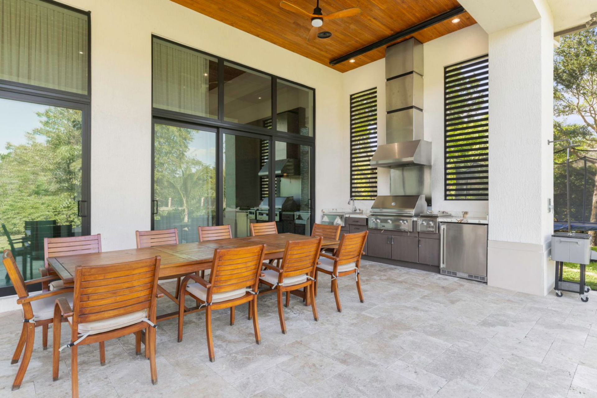 Beneath the wood-paneled ceiling, a full summer kitchen features a built-in grill, stainless vent hood, and refrigerator, positioned beside a long dining table that seats a large group for meals outdoors.
