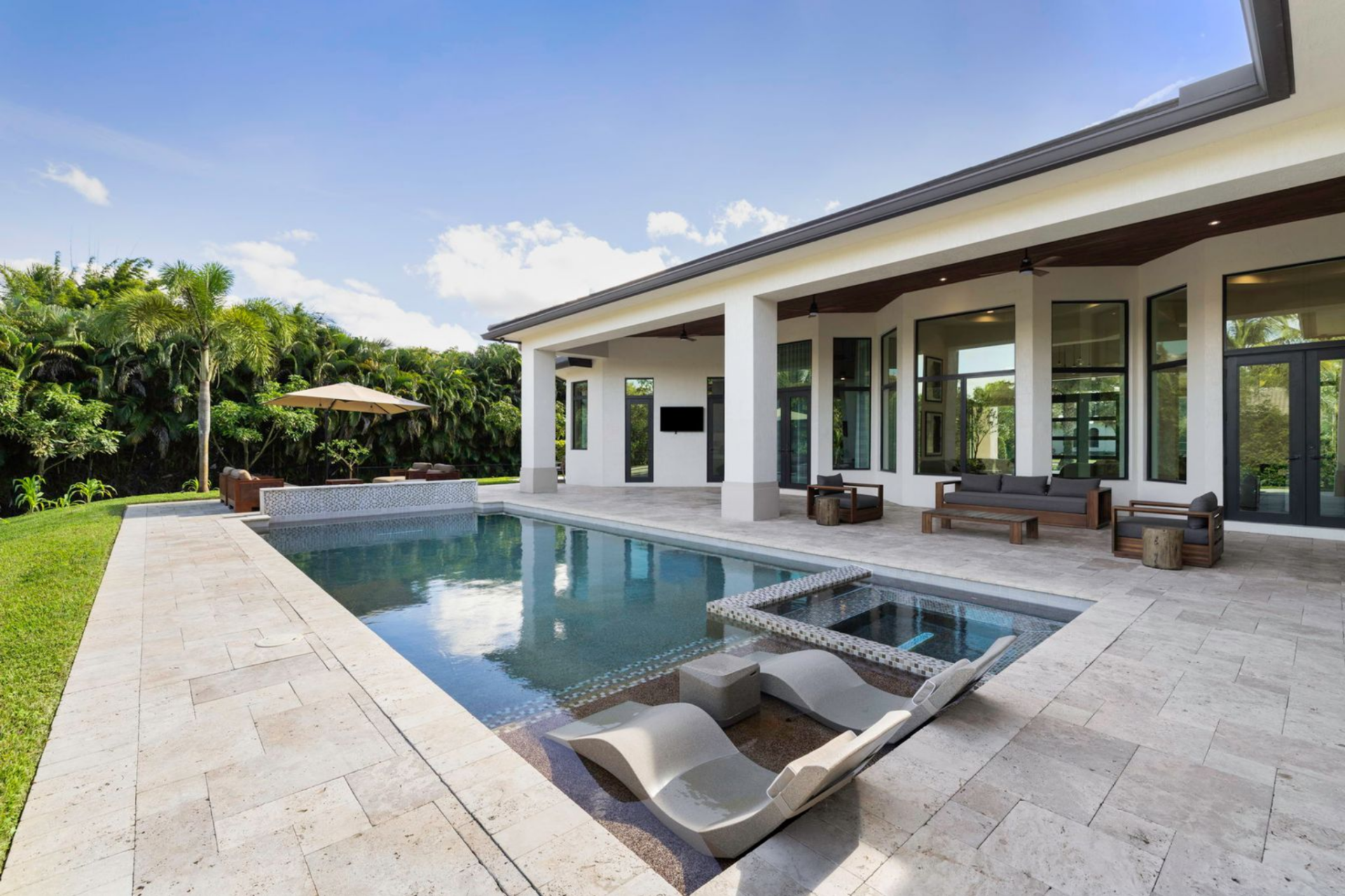 A shallow sun shelf with built-in loungers sits at one end of the pool, while additional seating areas are arranged beneath the covered terrace and along the surrounding deck.