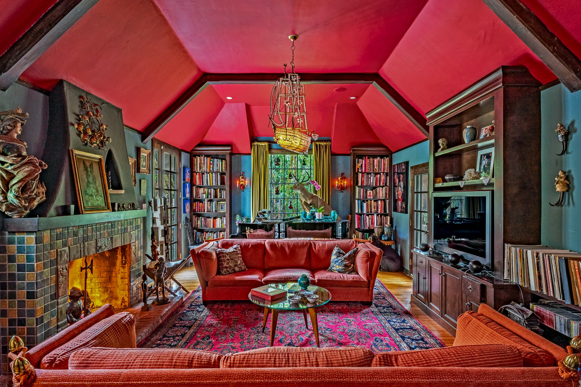 At the center of the space, a seating area built around patterned rugs and velvet sofas faces a tiled fireplace, while built-in shelves filled with books and artwork line the walls.