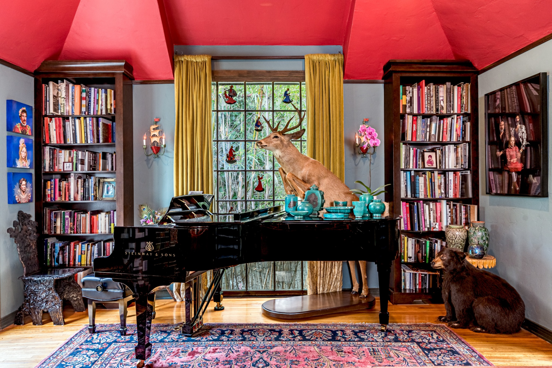 Toward the back of the room, a grand piano sits beneath a tall window framed with yellow curtains and stained-glass window accents, allowing natural light to filter in while highlighting the room’s eclectic mix of textures, color, and collected pieces.