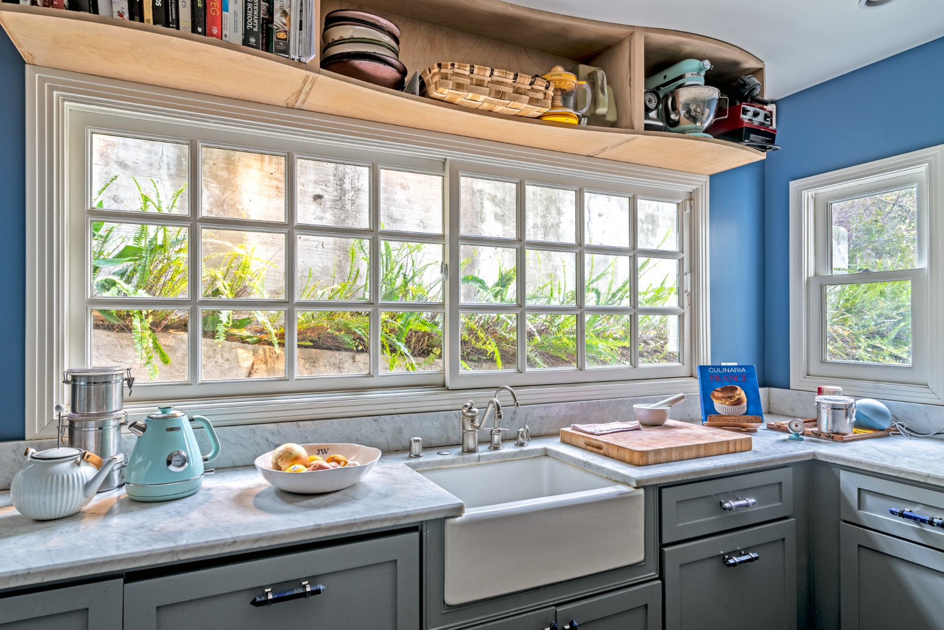 A farmhouse sink sits beneath a wide grid of windows that look out toward greenery just beyond the glass, bringing natural light into the workspace while open shelving above displays cookbooks, baskets, and small appliances.