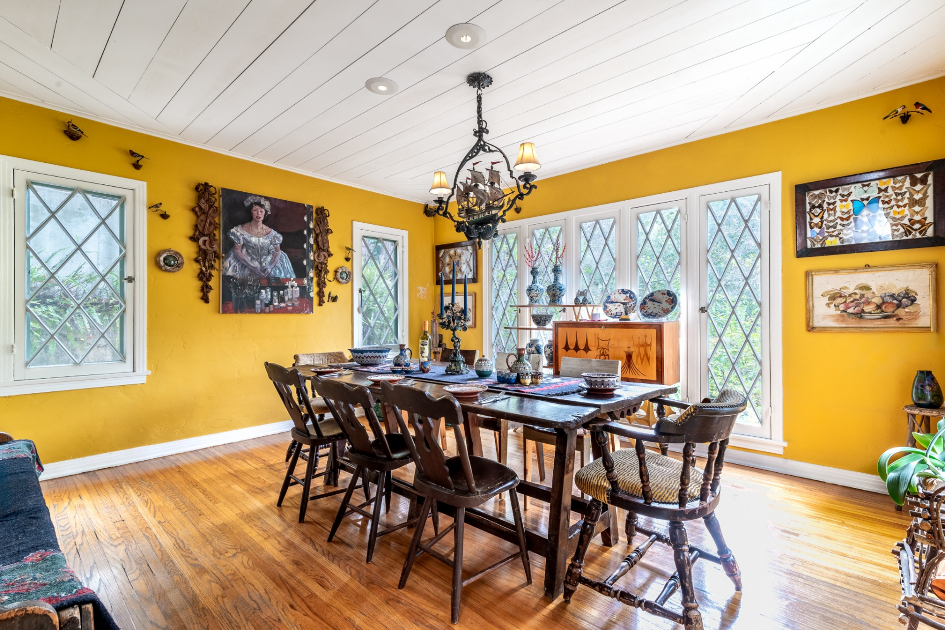 A long wooden dining table sits at the center beneath a decorative chandelier, surrounded by classic wooden chairs and set against hardwood floors that run throughout the room.