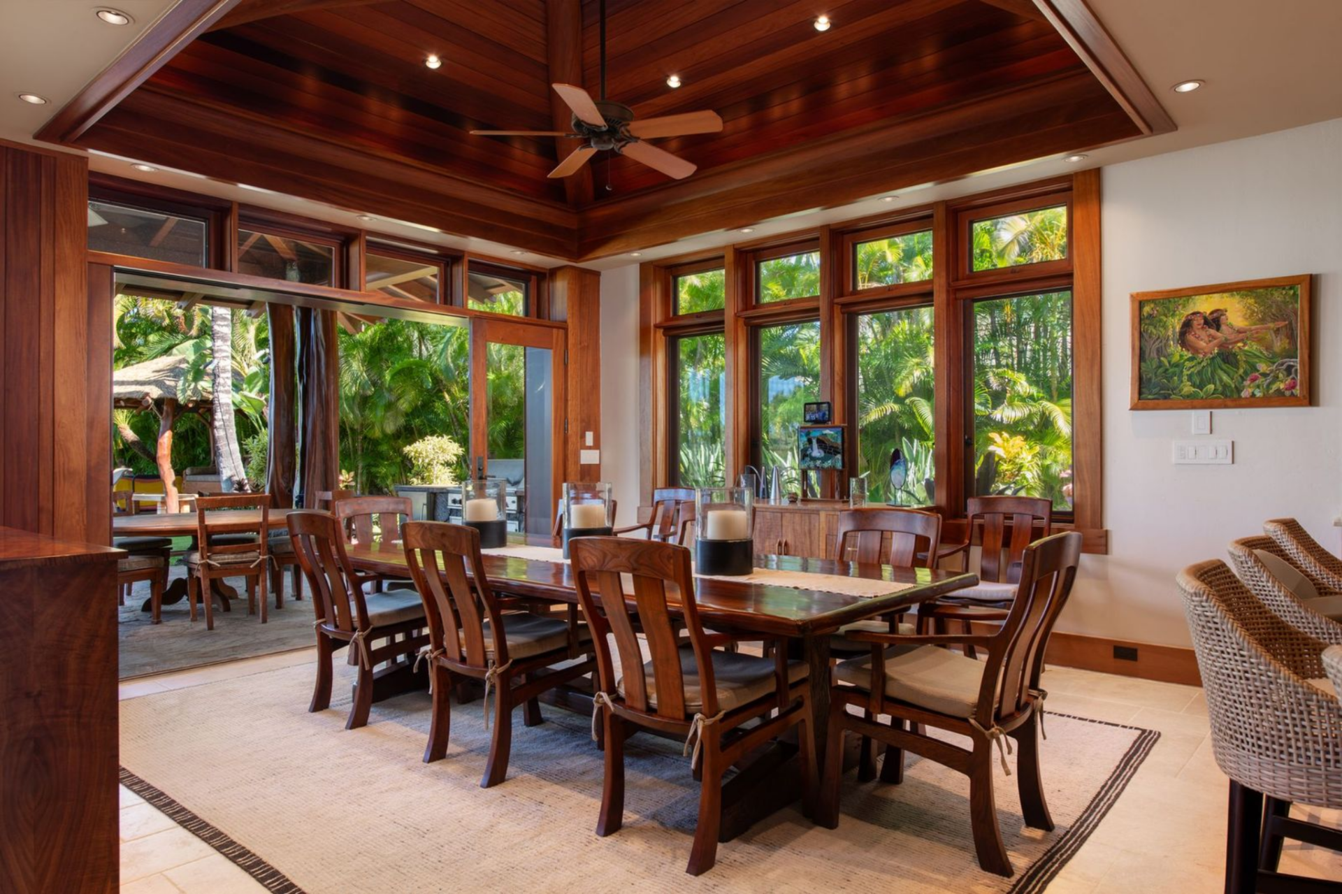 Around the corner from the great room, the formal dining room features a long wooden dining table surrounded by large windows that wrap the walls and frame views of the surrounding tropical landscaping.
