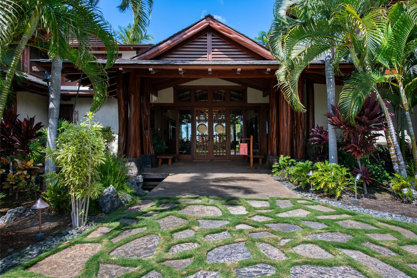 The front entrance itself is set beneath a wide overhang supported by massive timber posts, opening to a stone-floored lanai with built-in seating and custom wooden doors detailed with decorative latticework and hanging gongs. 