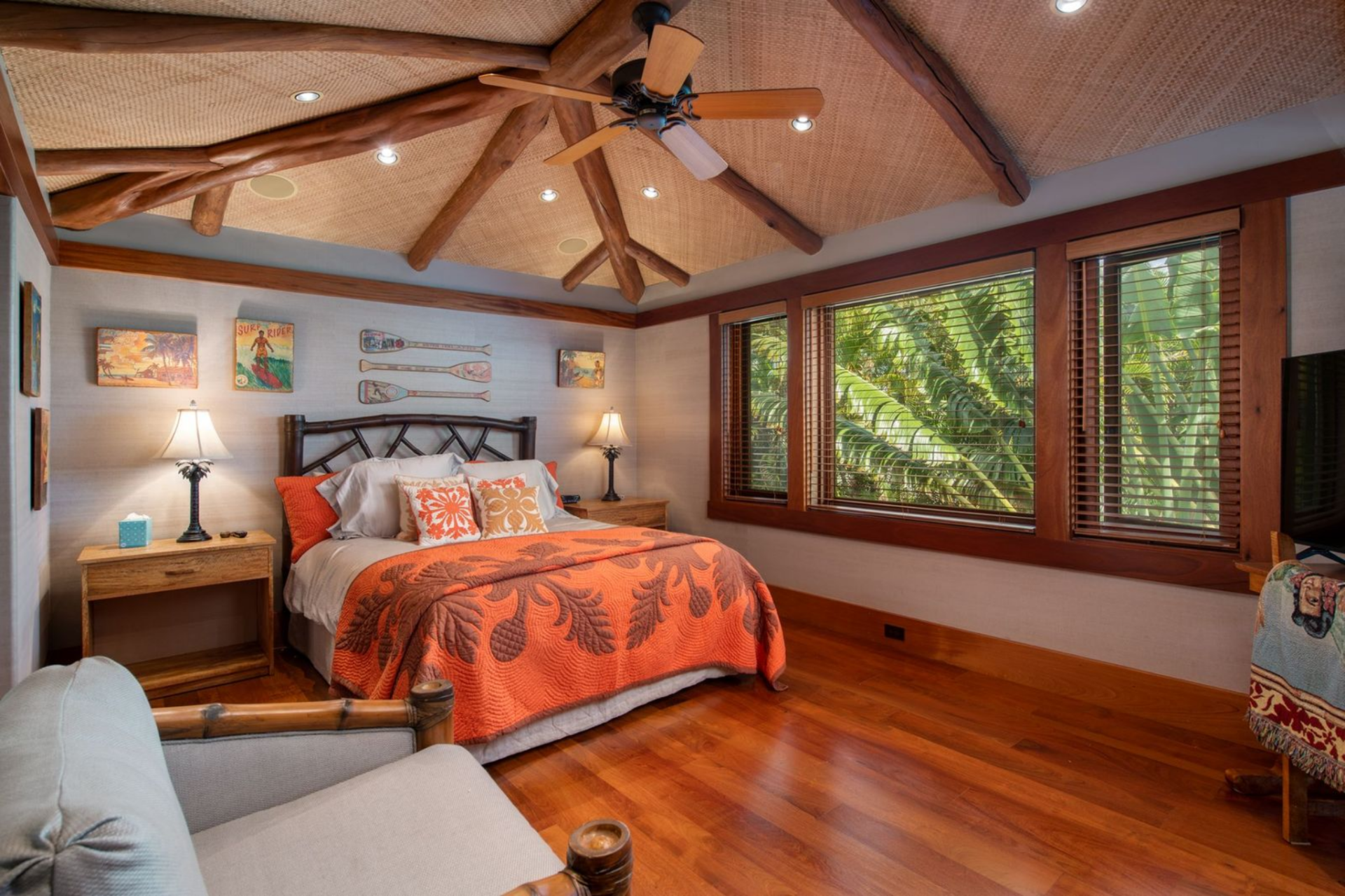 Another bedroom features a wood-framed bed with colorful bedding that adds contrast against the warm structure of the house and a row of wide windows frames tropical plants just outside.