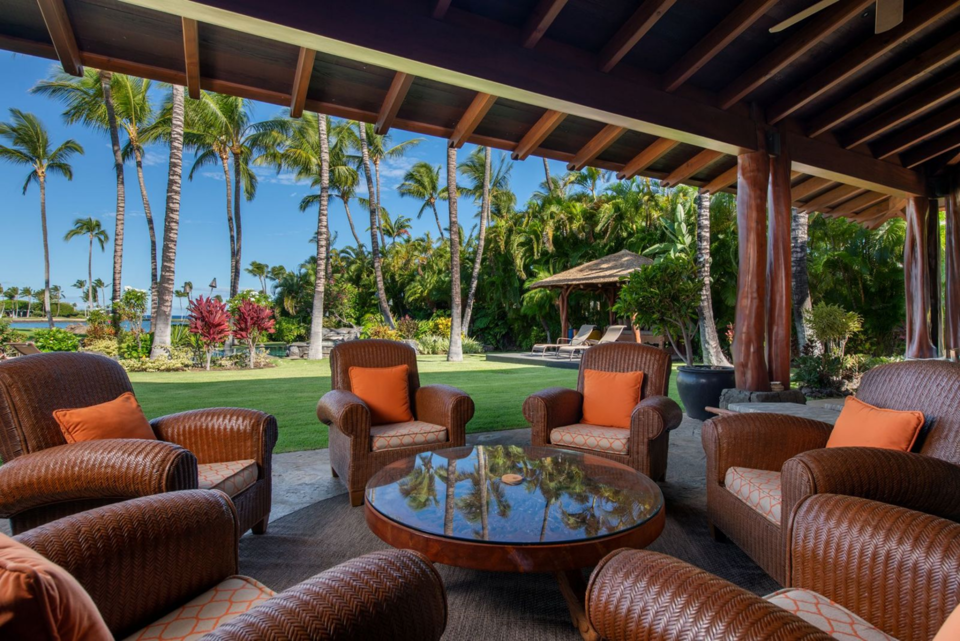 Large timber posts support the deep overhangs while seating areas arranged with wood-framed sofas and wicker chairs create multiple gathering spots that look out across the landscaped grounds.
