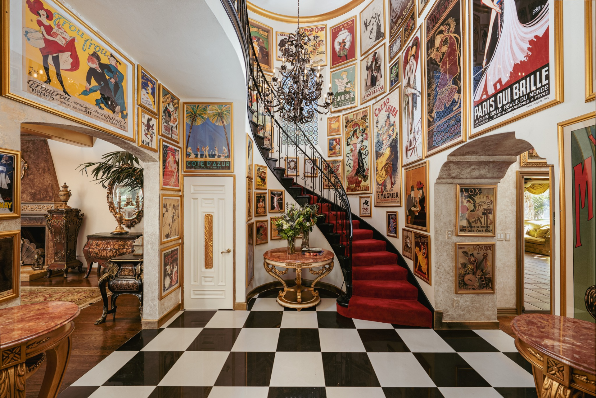 A sweeping curved staircase with a red carpet runner rises along the wall, framed by a wrought-iron railing and crowned by a chandelier that hangs above the black-and-white checkered marble floor.