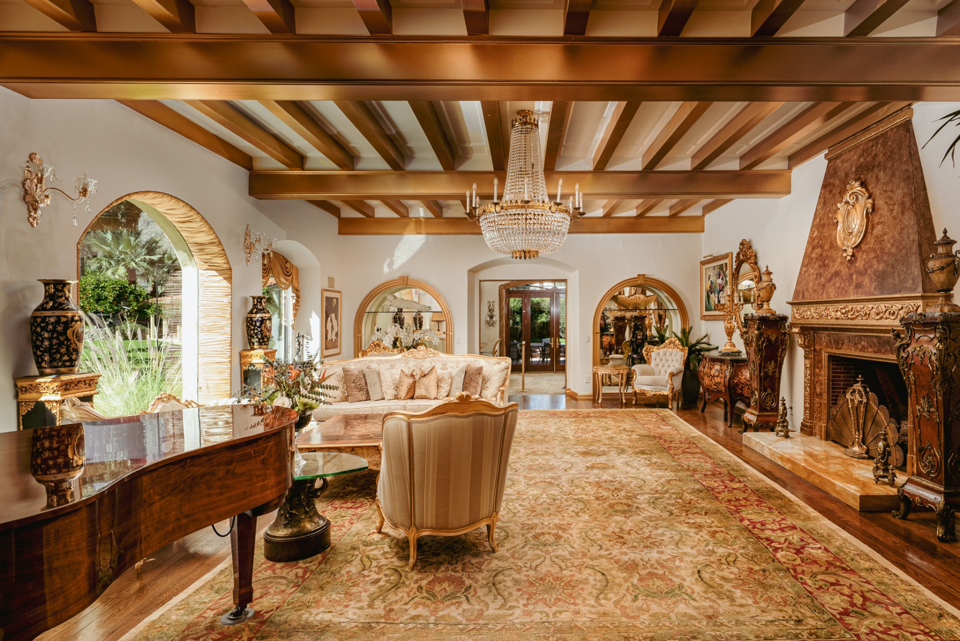 Arched windows draw in natural light and frame views of the surrounding greenery, while a carved fireplace with an elaborate mantel provides a focal point along one wall. 
