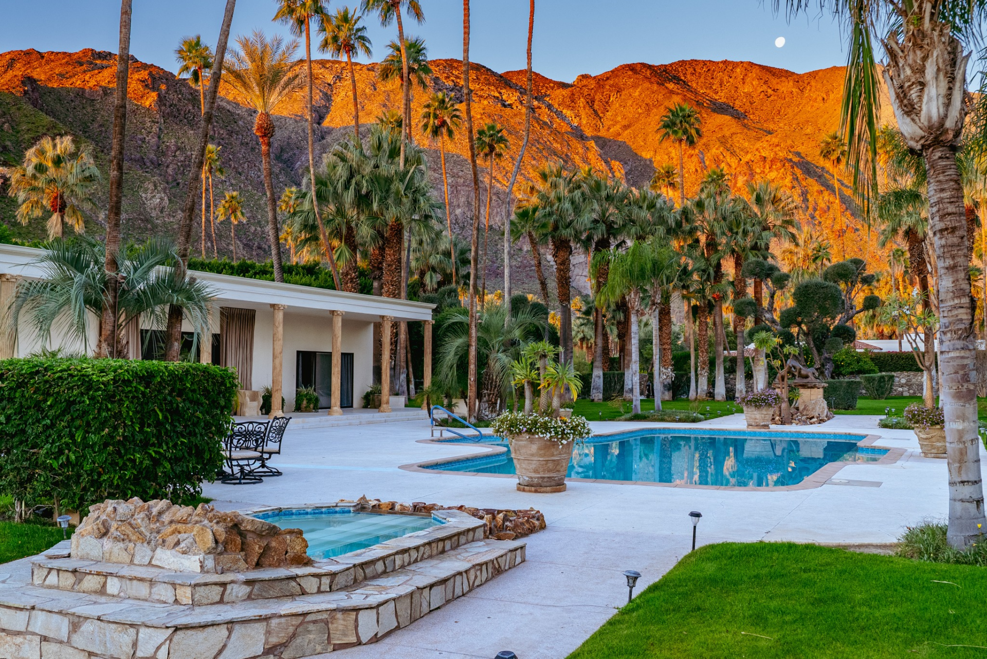 A raised spa sits beside the pool, while expansive patios and manicured lawns provide plenty of space for outdoor seating and lounging beneath the towering palms.