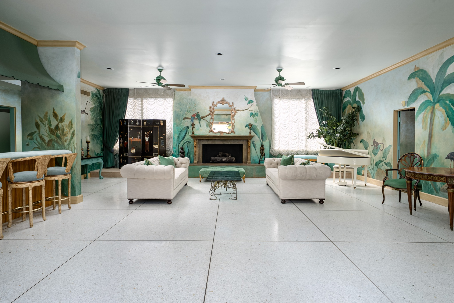 Behind the pool, the pool house functions as a full secondary gathering space with interiors wrapped in hand-painted tropical murals depicting palms, birds, and lush greenery.