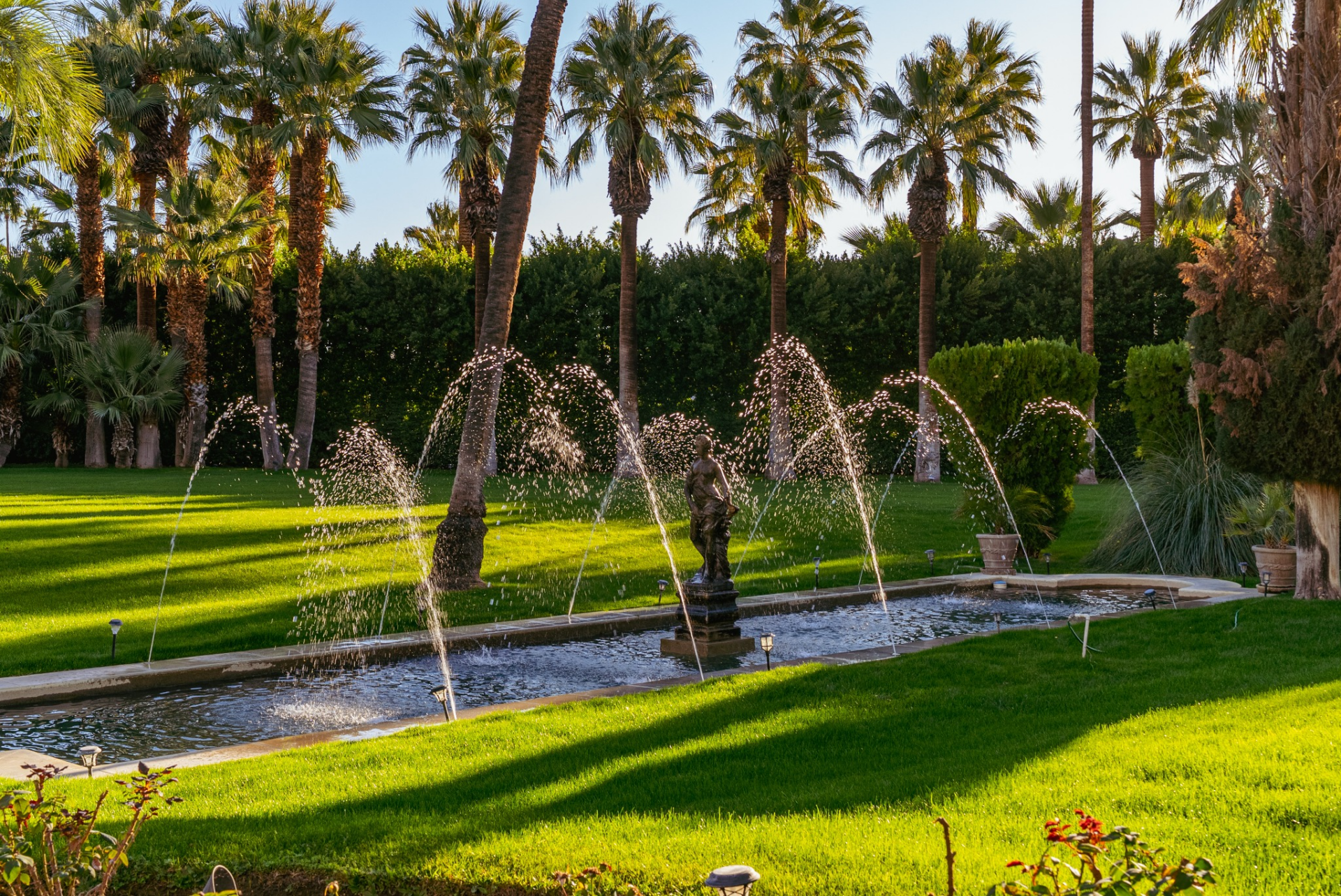 A long reflecting fountain runs through one portion of the property, where sculpted water jets arc over a central statue, creating a peaceful water element framed by rows of plants.