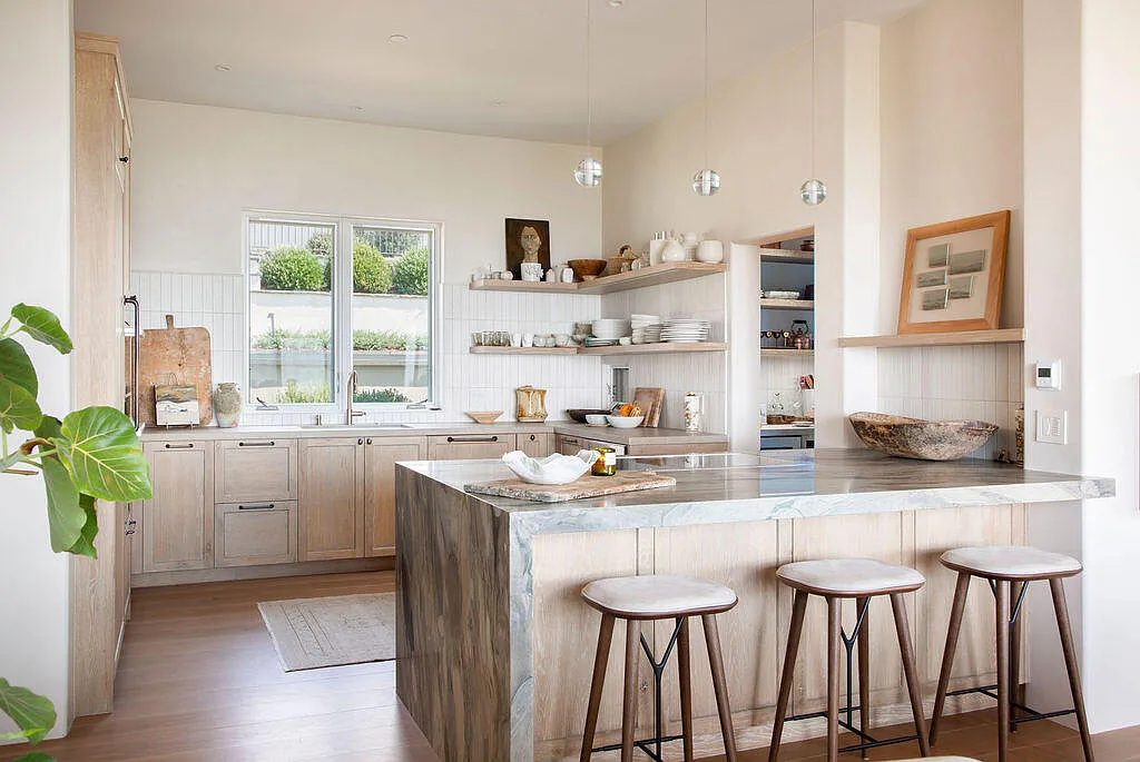 Light wood cabinetry and open shelving displaying everyday dishware line the walls, while simple pendant lights hang above the island, creating a bright and cozy design.