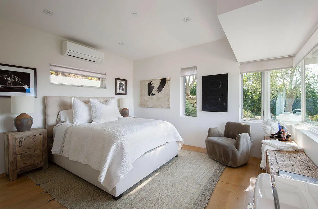 The guest house's bedroom has large windows that wrap the room, bringing in natural light and views of the surrounding greenery. 