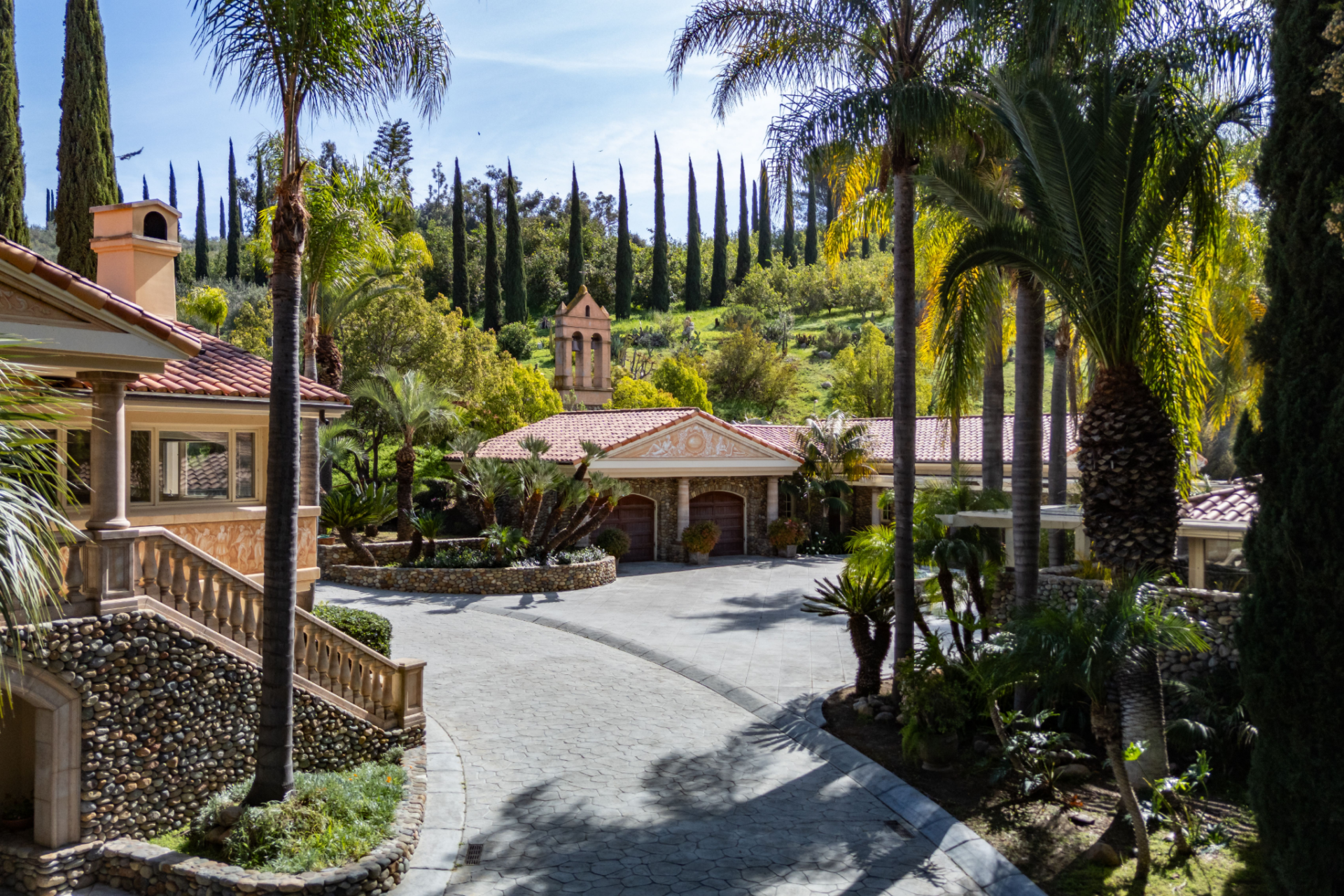 Positioned among palms and Mediterranean-style landscaping, the residence sits behind a broad motor court, with a tiled roof, stone accents, and arched architectural details.