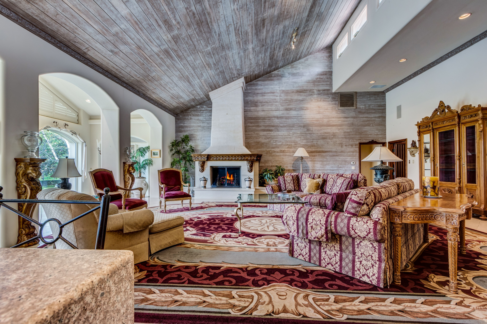 At the center of the room, a tall white fireplace rises to the peak of the ceiling, framed by a carved mantel across from patterned sofas, carved wood tables, and antique display cabinets.