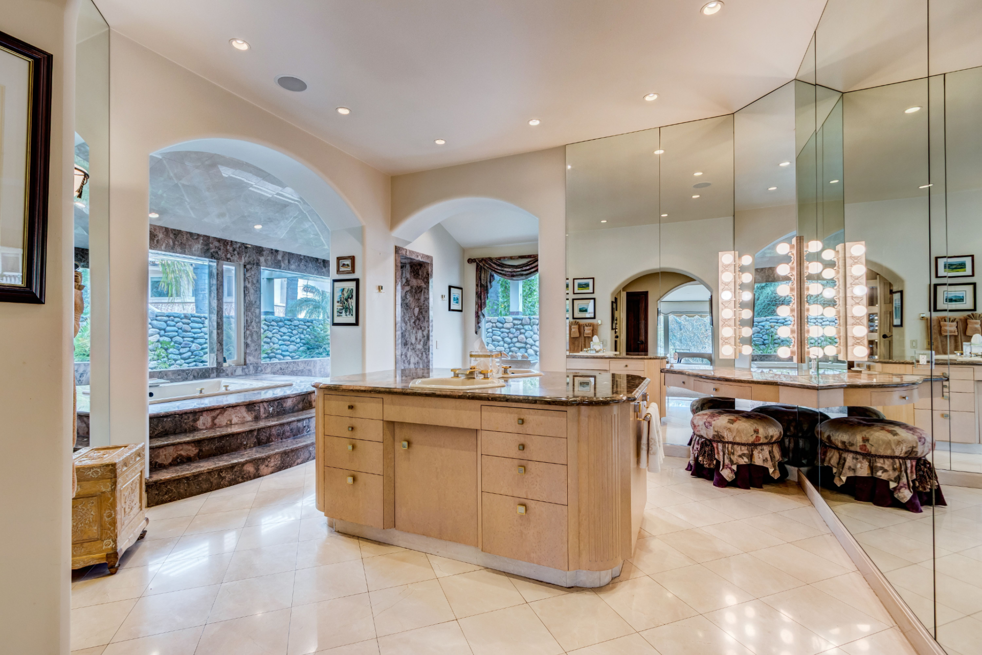 A mirrored wall with theater-style lighting surrounds the vanity area, while arched openings lead deeper into the bathroom, where a large soaking tub is set into a raised marble. 