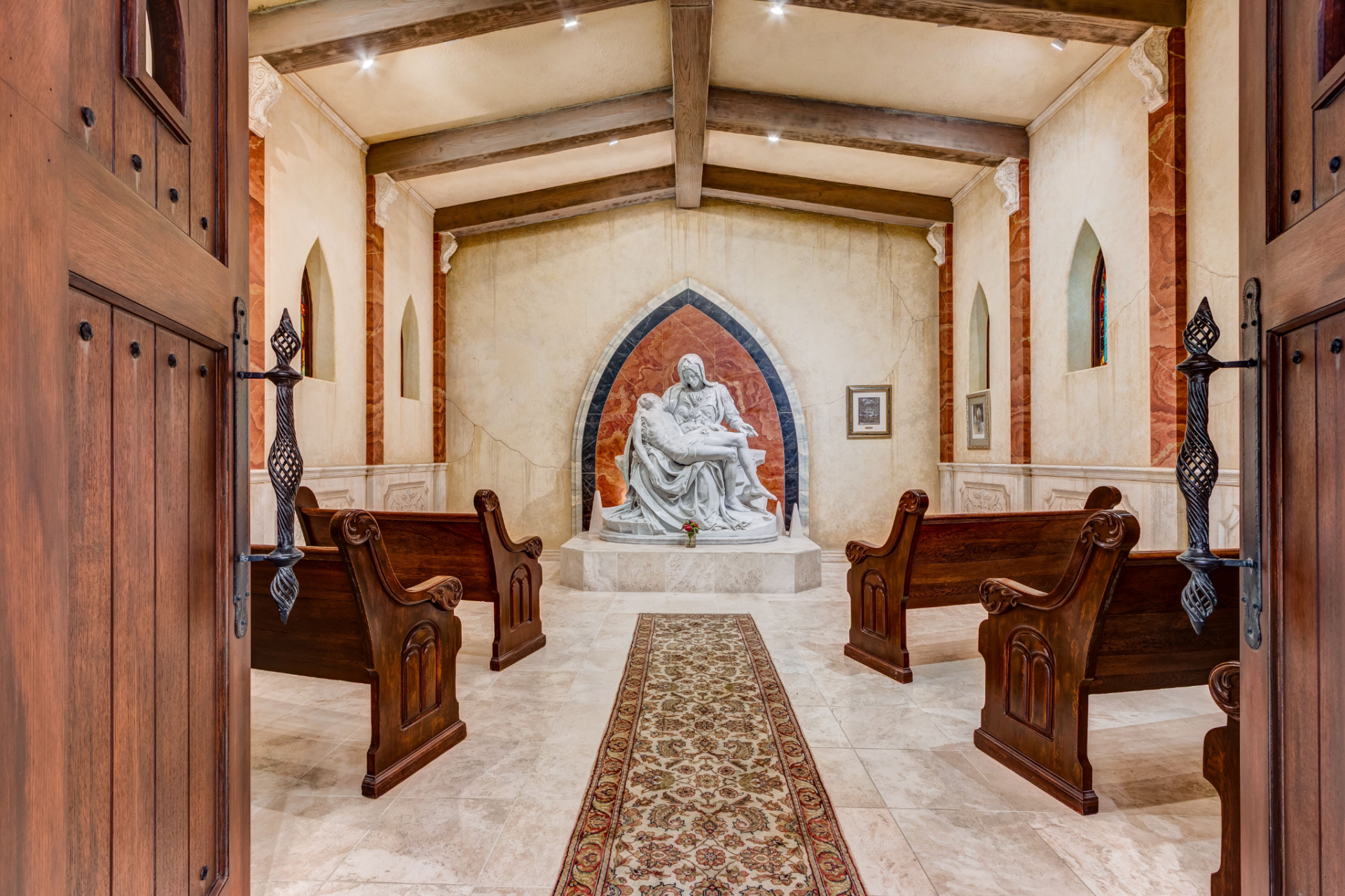 Entered through heavy wooden doors, the room features rows of carved wooden pews facing a marble sculpture set within an arched alcove framed by rich stone detailing. 
