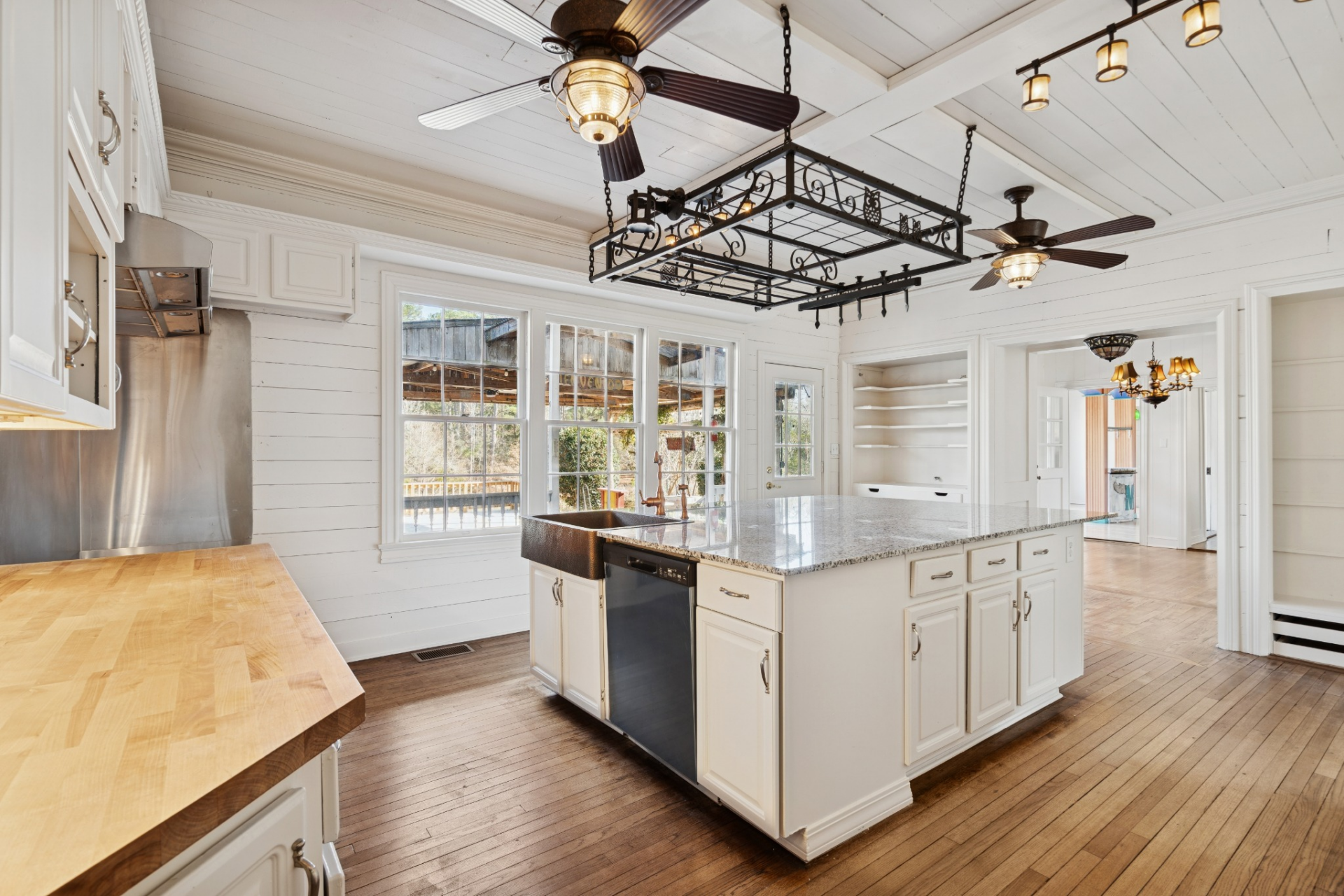 Natural light fills the room through a series of windows overlooking the exterior, while stainless steel appliances and butcher block counters add contrast against the lighter palette. 
