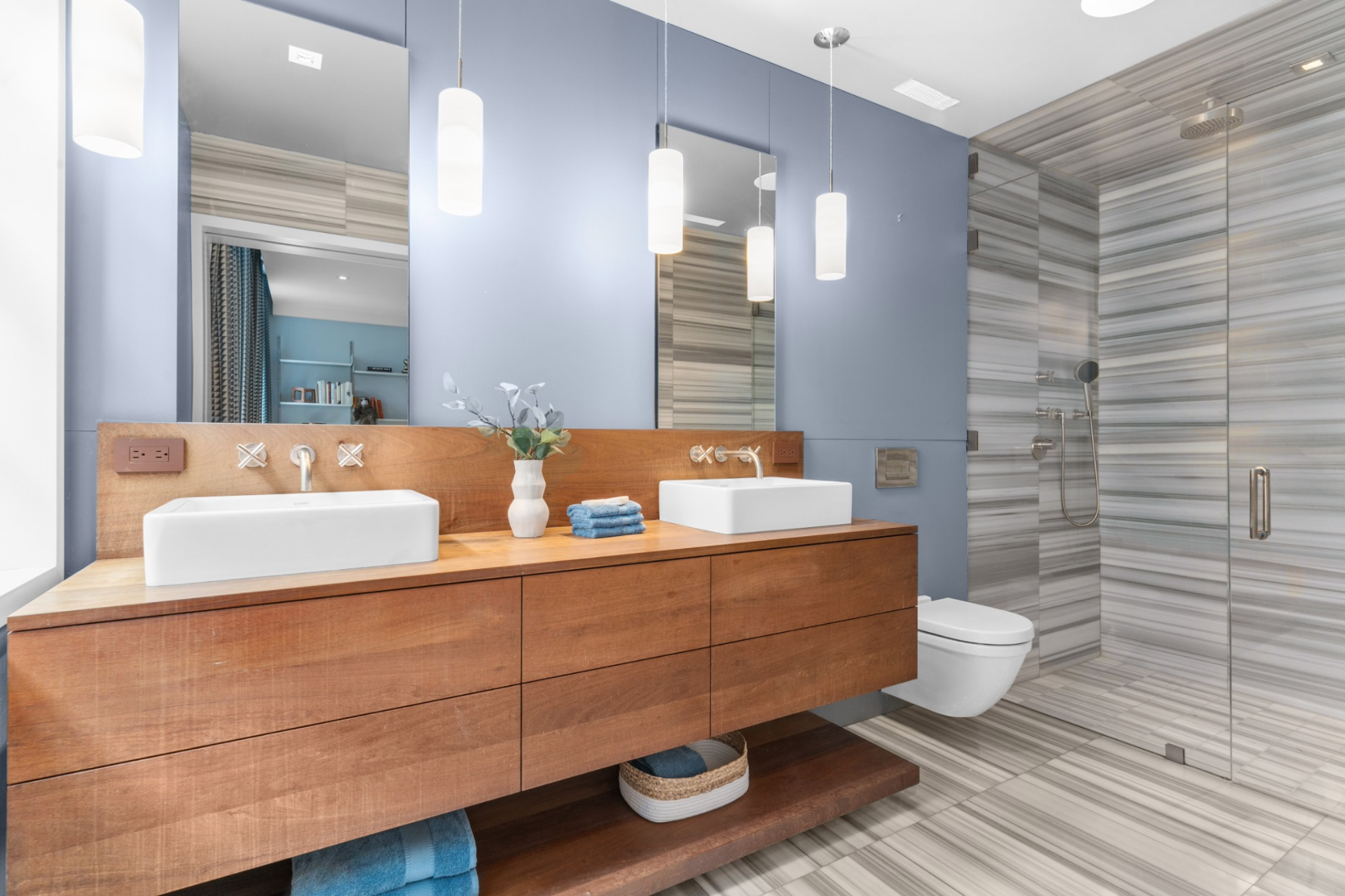 Its bathroom mirrors the home’s contemporary style, featuring a dual-sink wood vanity, modern lighting, and a glass shower finished with horizontal tile that extends to the floor for a more textured look.