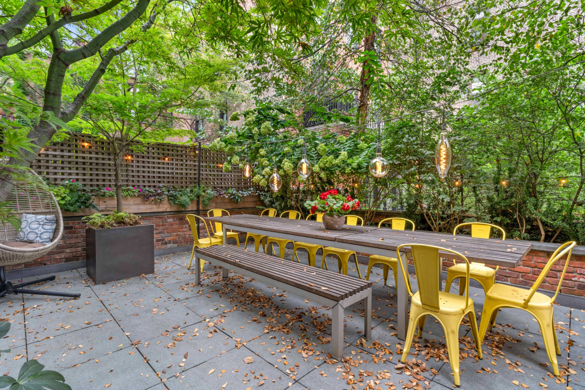 A long dining table sits beneath string lighting, creating a defined space for larger gatherings, while planters and trees soften the edges of the layout.
