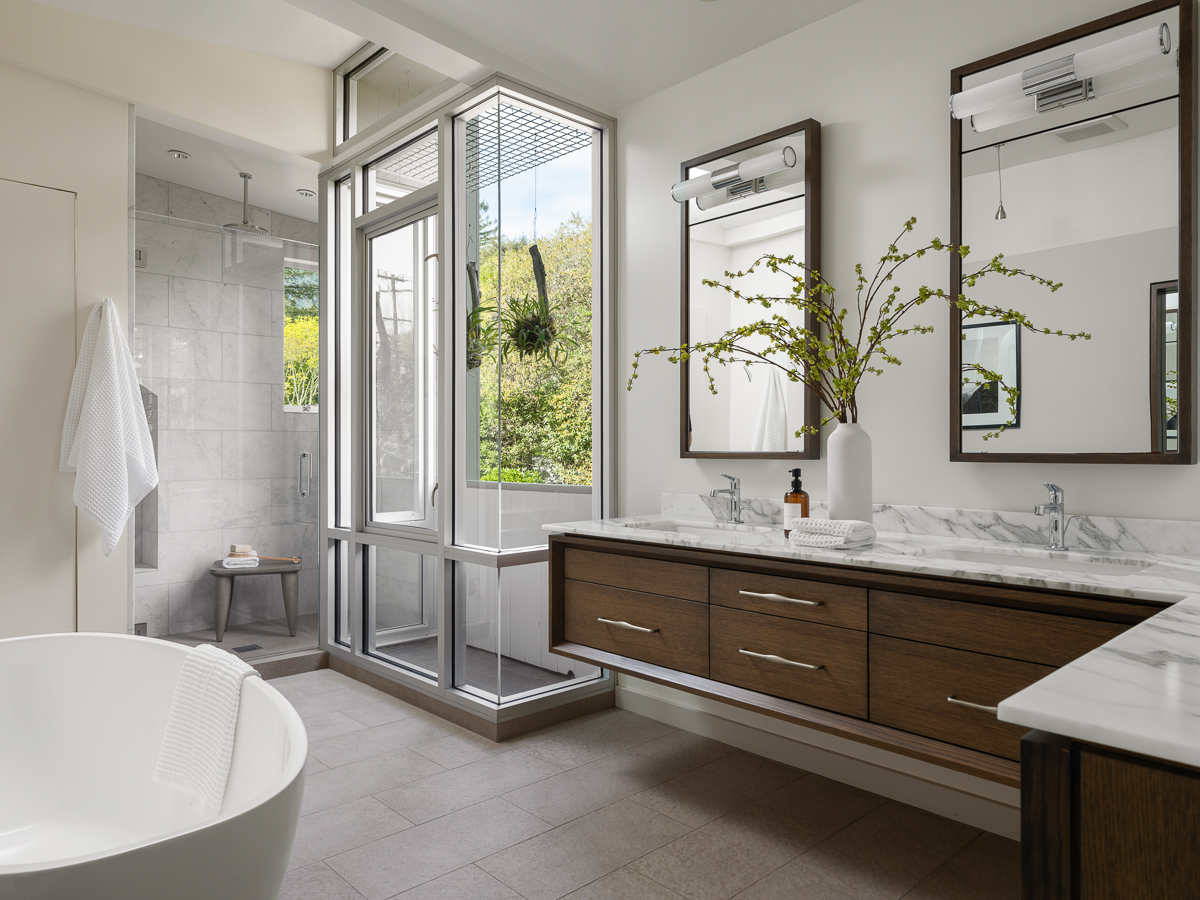 The en-suite bathroom features a double vanity with marble countertops that runs along two walls, while a glass-enclosed shower sits beside a windowed corner that brings in light and greenery. 