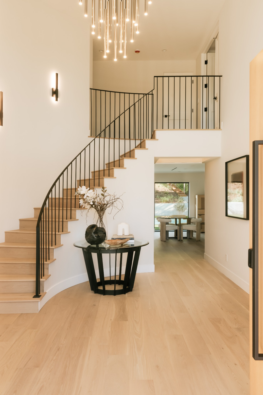 A double-height ceiling draws the eye upward, where a cascading light fixture hangs above a curved staircase with dark metal railing and light wood treads.