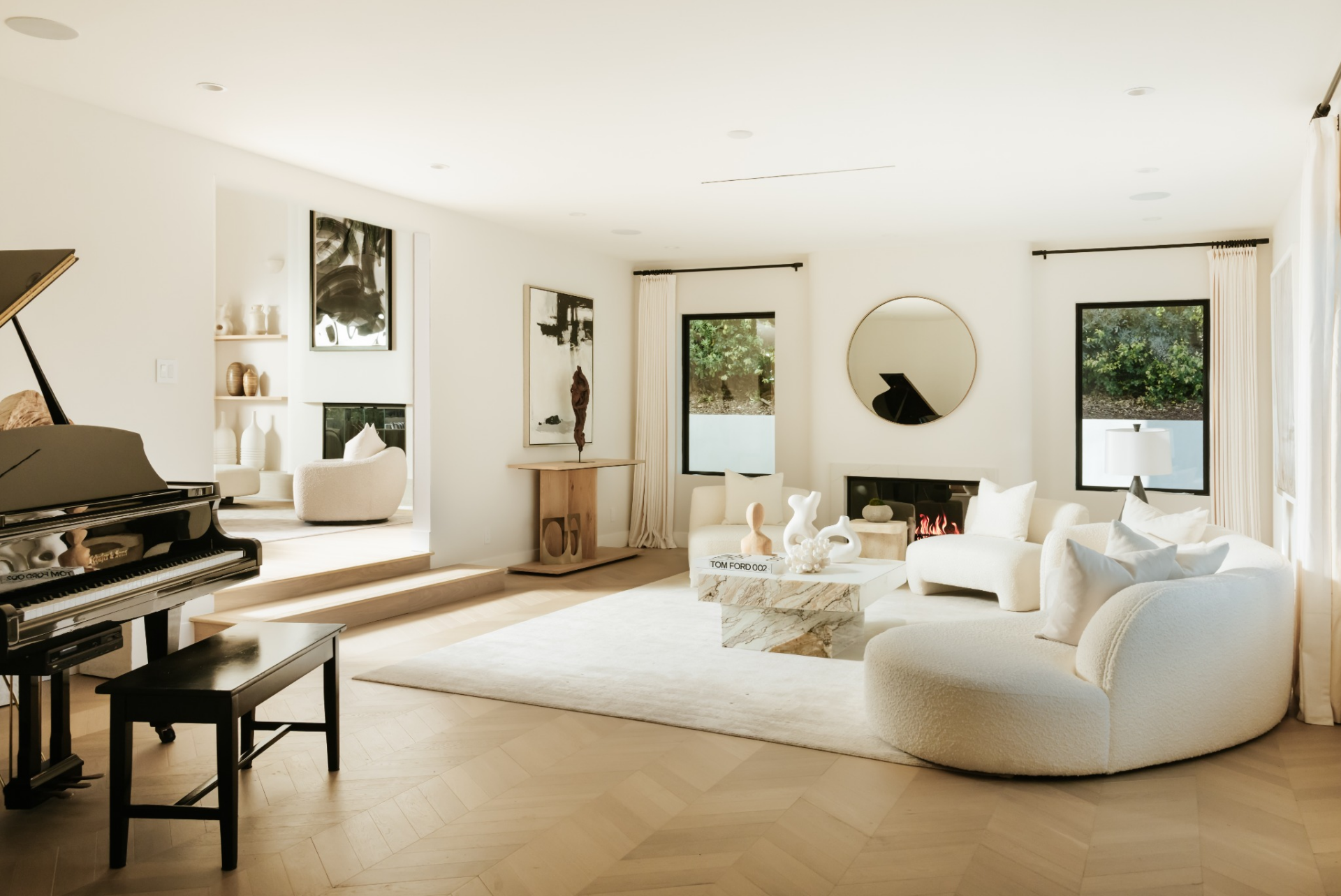 A fireplace sits on one wall beneath a round mirror, while large windows on either side bring in natural light reflecting off a grand piano in the other corner.