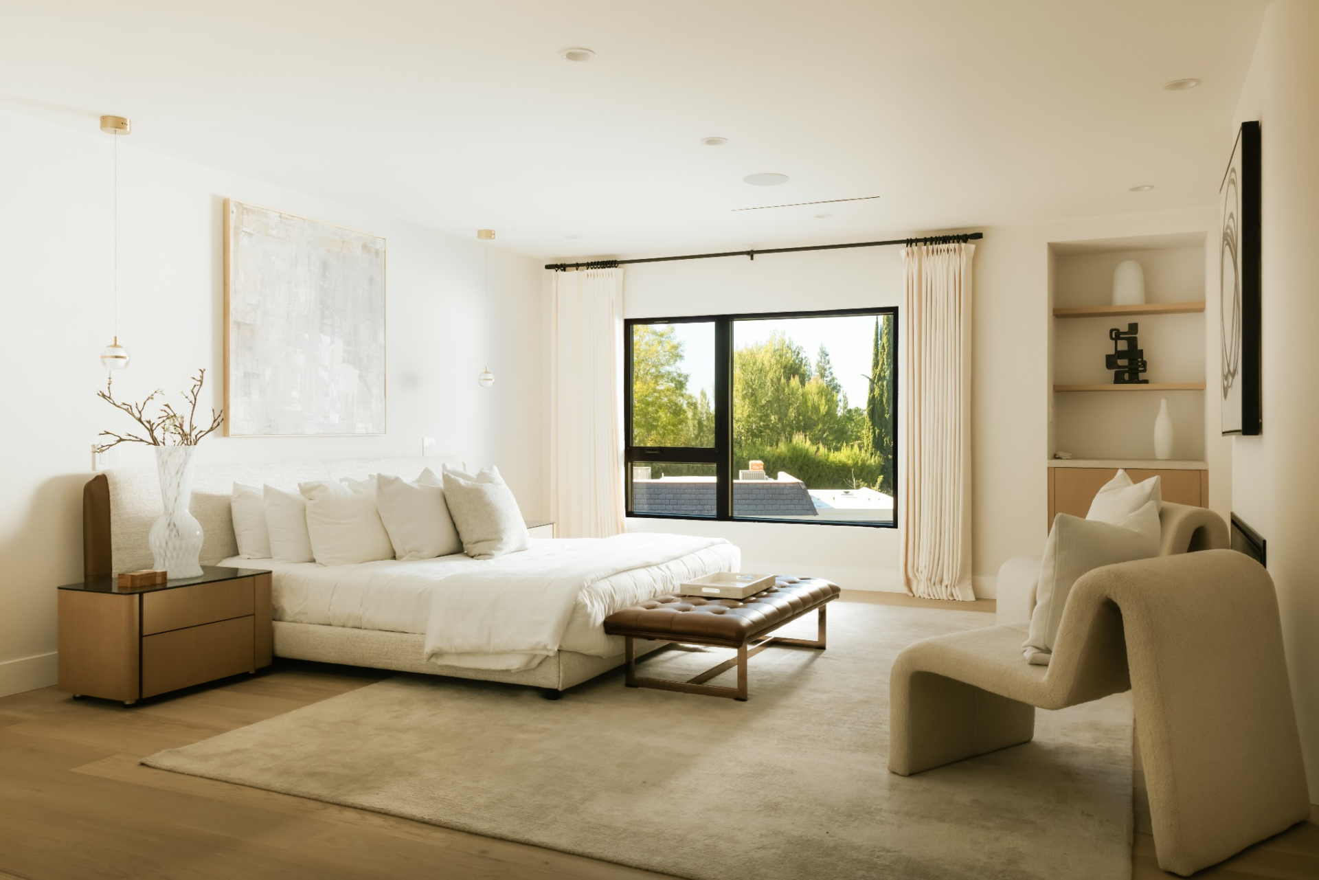 The bedroom centers around a low-profile upholstered bed set against a soft neutral backdrop, with wide-plank flooring continuing across the ground. 