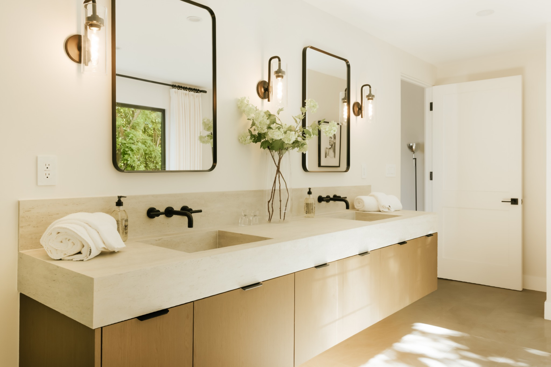 The double vanity runs along the final wall with integrated sinks, flat-panel cabinetry, and black hardware, topped by framed mirrors and wall-mounted lighting.