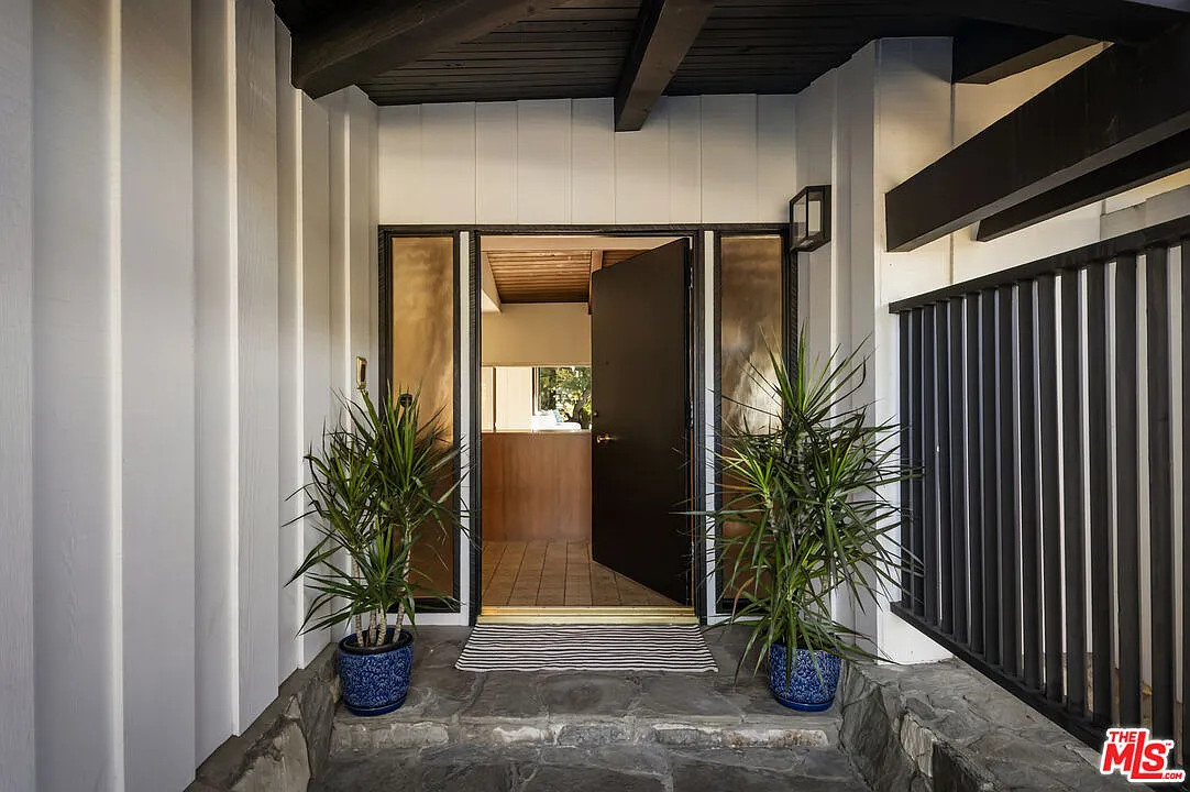 A covered front porch with exposed beams leads to a simple, dark-framed doorway framed by narrow glass panels.