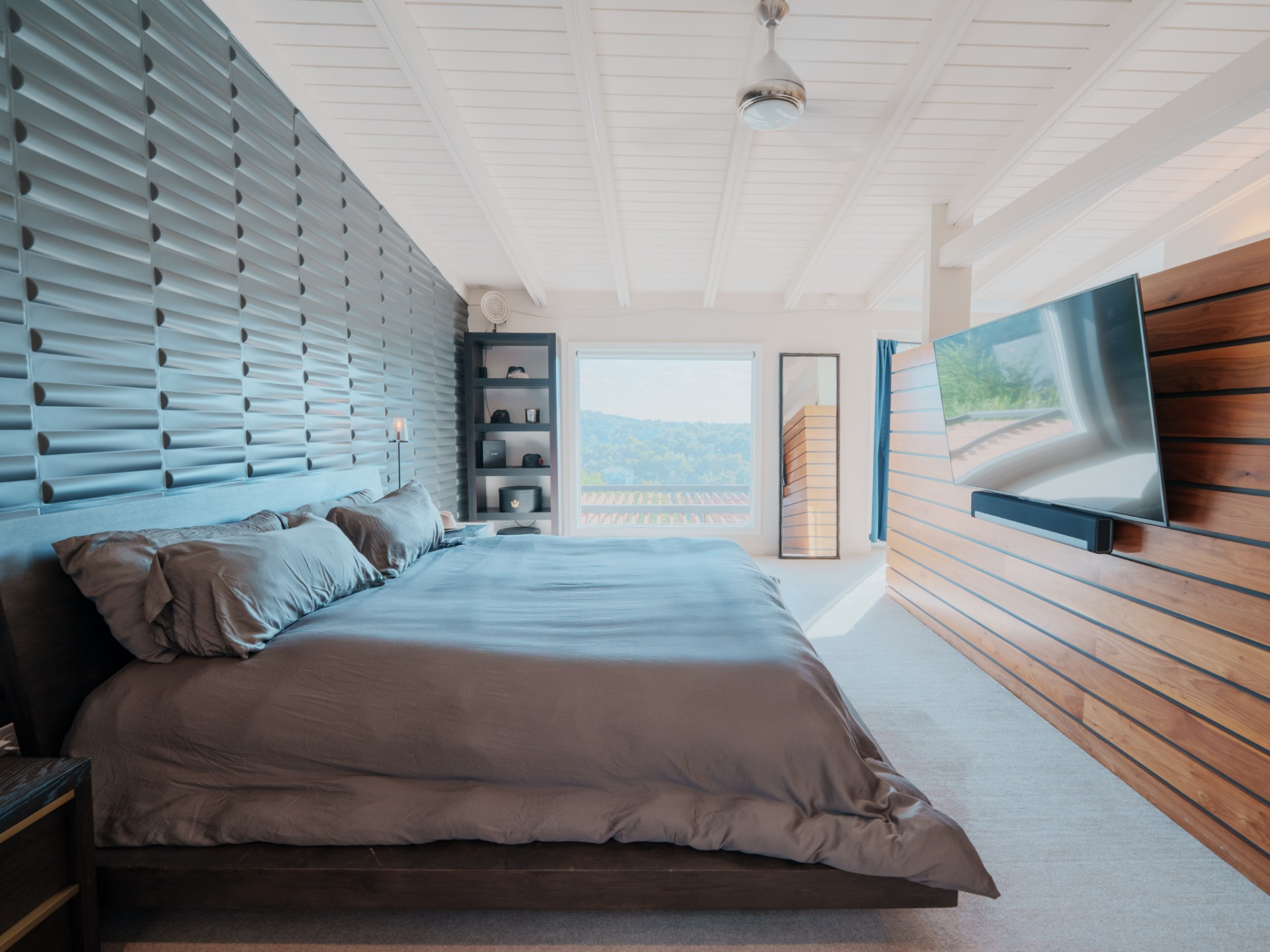 Across from it, horizontal wood slats anchor a mounted television and soundbar, bringing warmth and contrast beneath the white beamed ceiling and modern ceiling fan overhead.