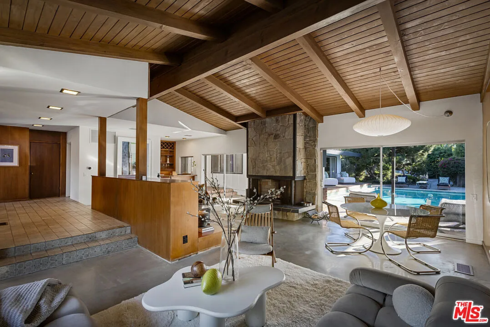 On the other side of the entrance, the home opens into a sunken great room, where distinct living zones unfold beneath exposed wood beams and extend toward the backyard through walls of glass.