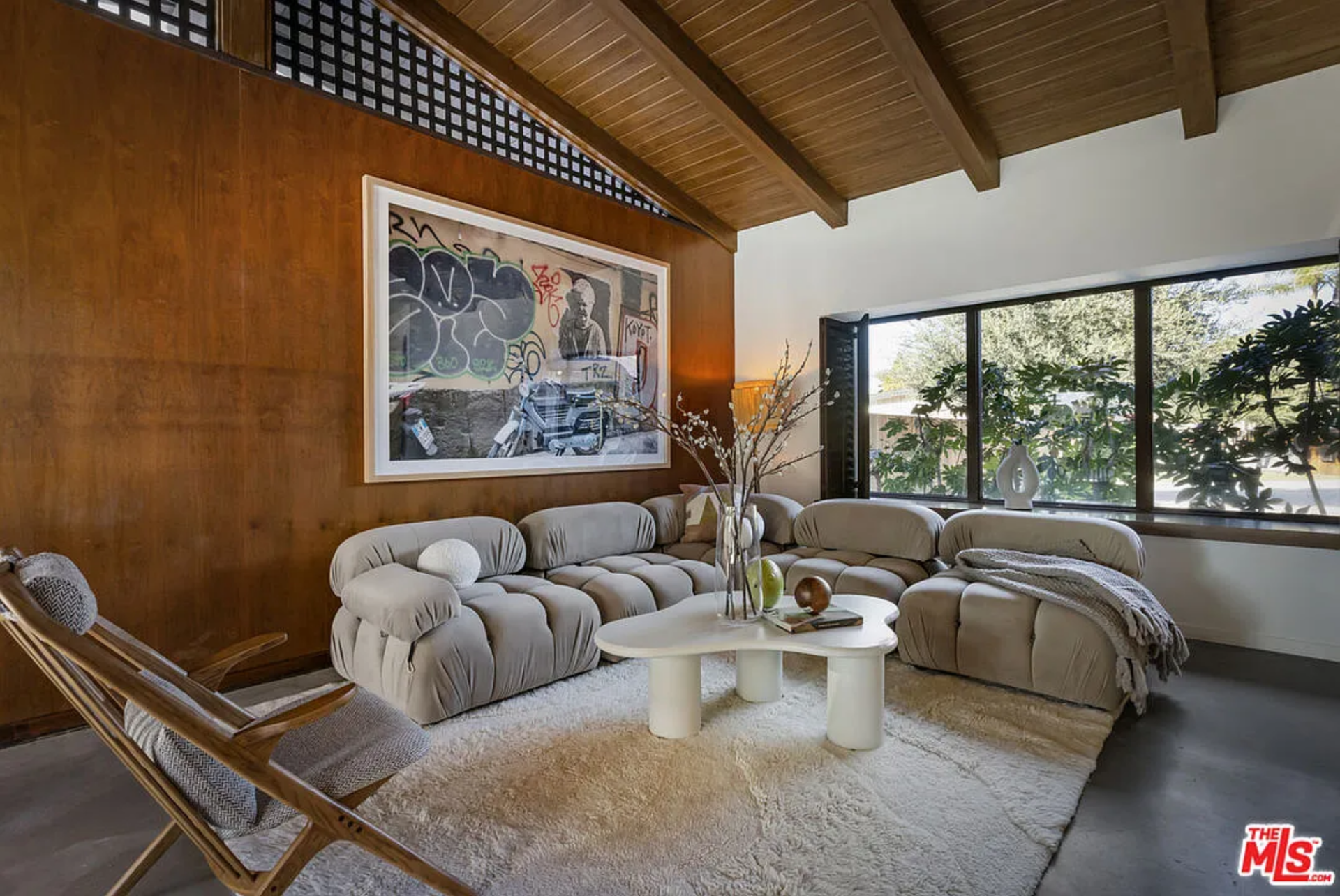 A low, sculptural sofa and lounge chair sit atop a textured rug, while a large window brings in natural light and greenery from the front of the property.