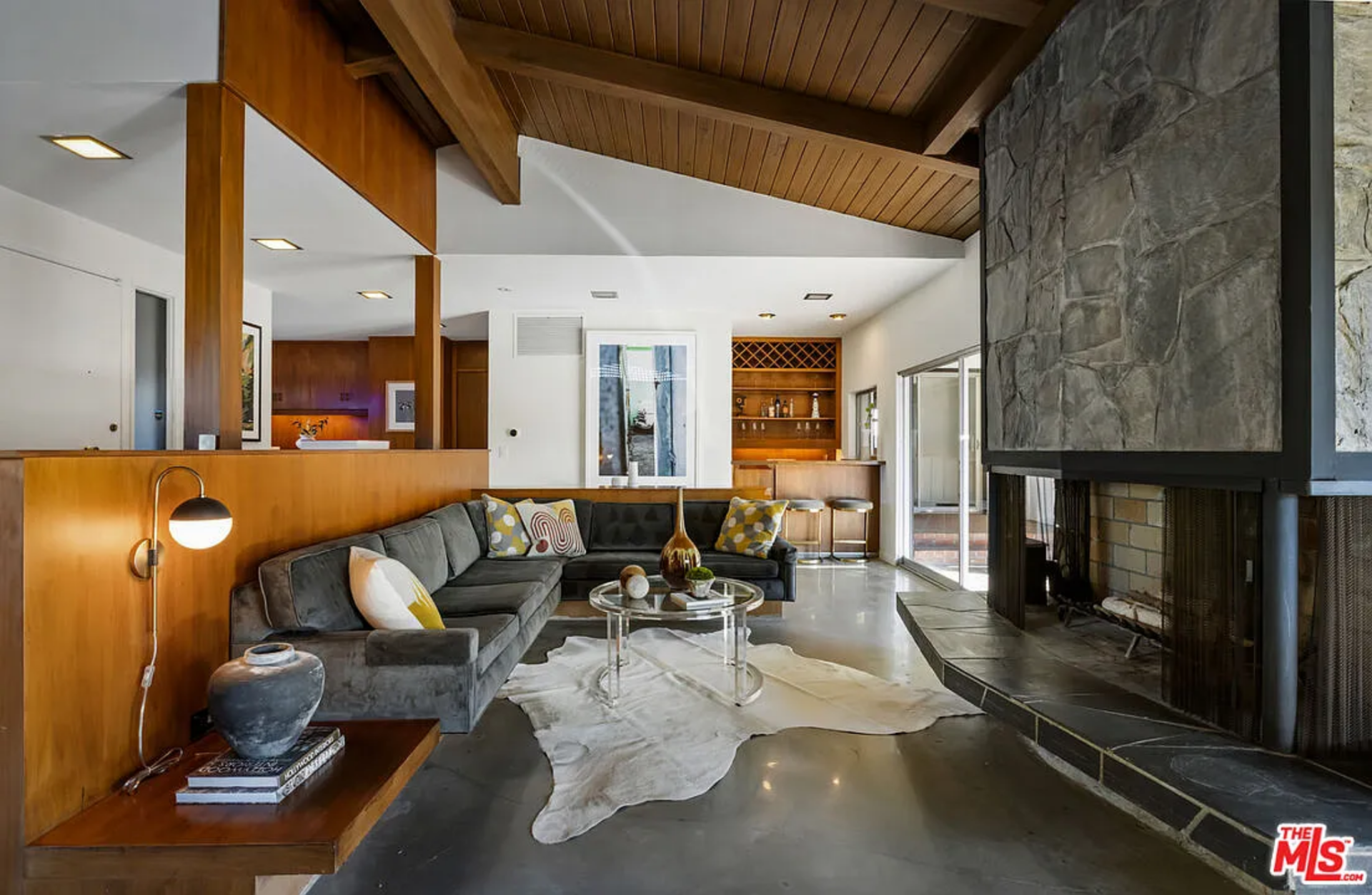 At the center, the sunken living area is defined by a sculptural stone fireplace that rises up to the ceiling, acting as both a focal point and a divider within the open plan. 