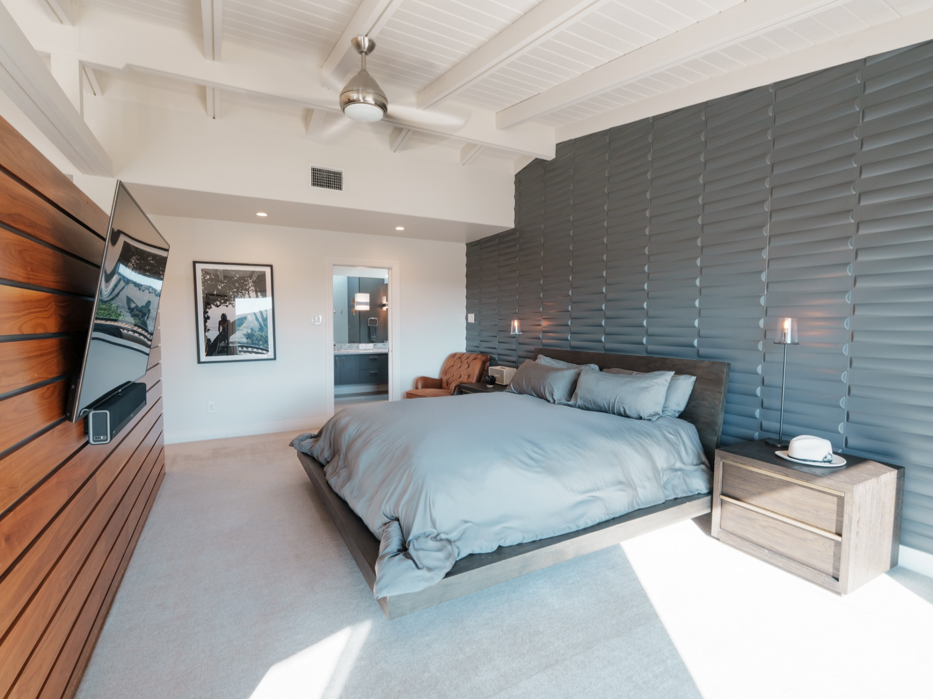 The suite begins with the bedroom, where a full wall behind the bed is wrapped in textured, sculptural paneling in a muted blue-gray. 