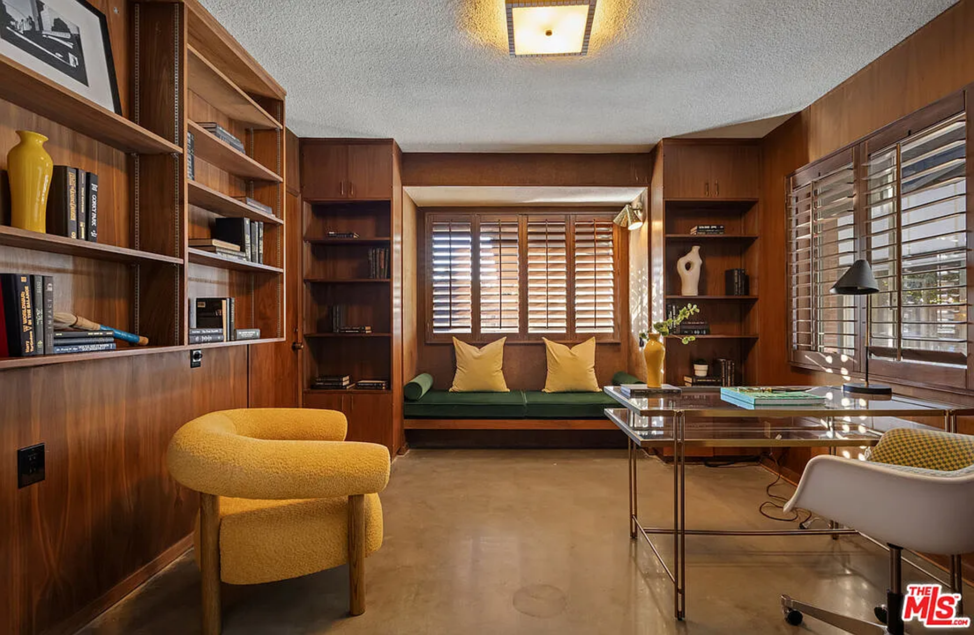 Wrapped in warm wood paneling, the office is lined with built-in shelving and anchored by a window seat tucked beneath plantation shutters. 