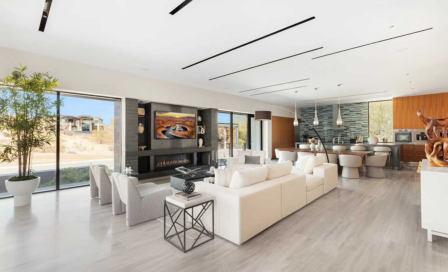 Floor-to-ceiling glass lines the walls, bringing in natural light and framing desert views, while the layout keeps the seating area centered around a long, linear fireplace set into a dark stone feature wall.