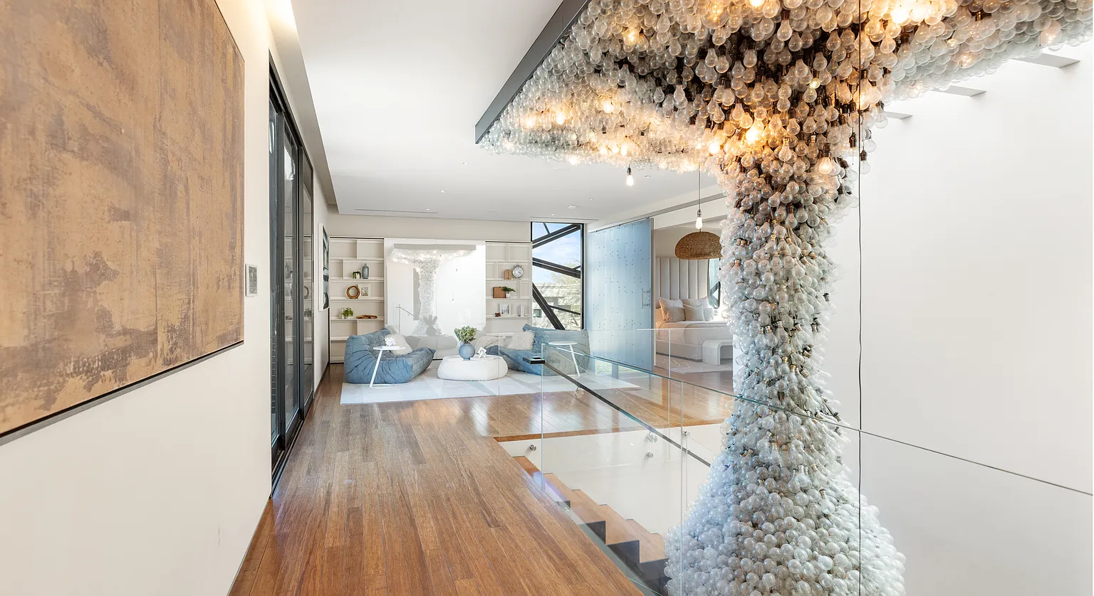 The area is positioned along a glass-lined landing, overlooking the level below. Overhead, a sculptural light installation made up of clustered lightbulbs turns the lounge into a statement space.