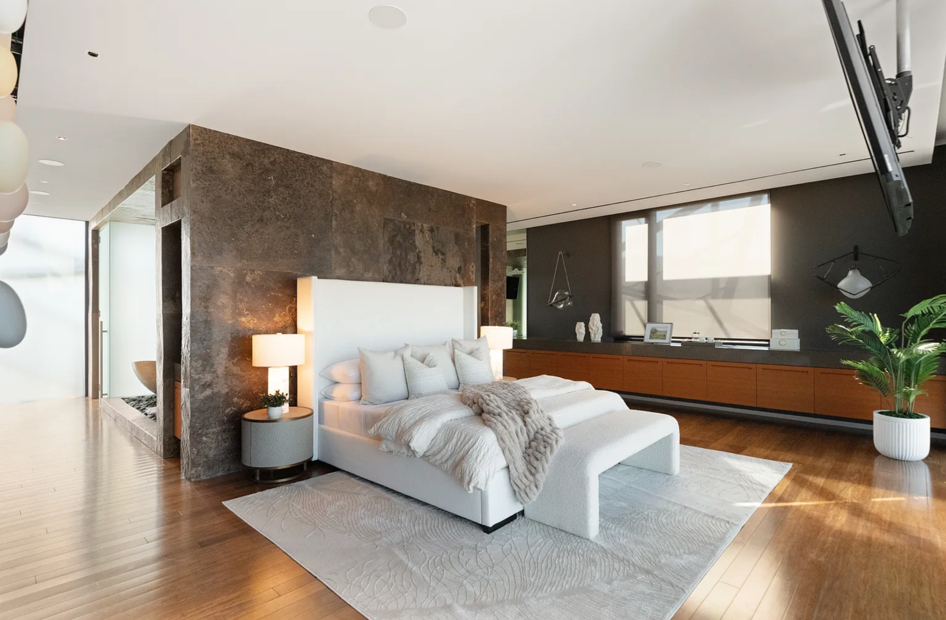 Inside, a large upholstered bed sits centered on a soft area rug, backed by a full-height stone wall that separates the sleeping area from the rest of the suite. 