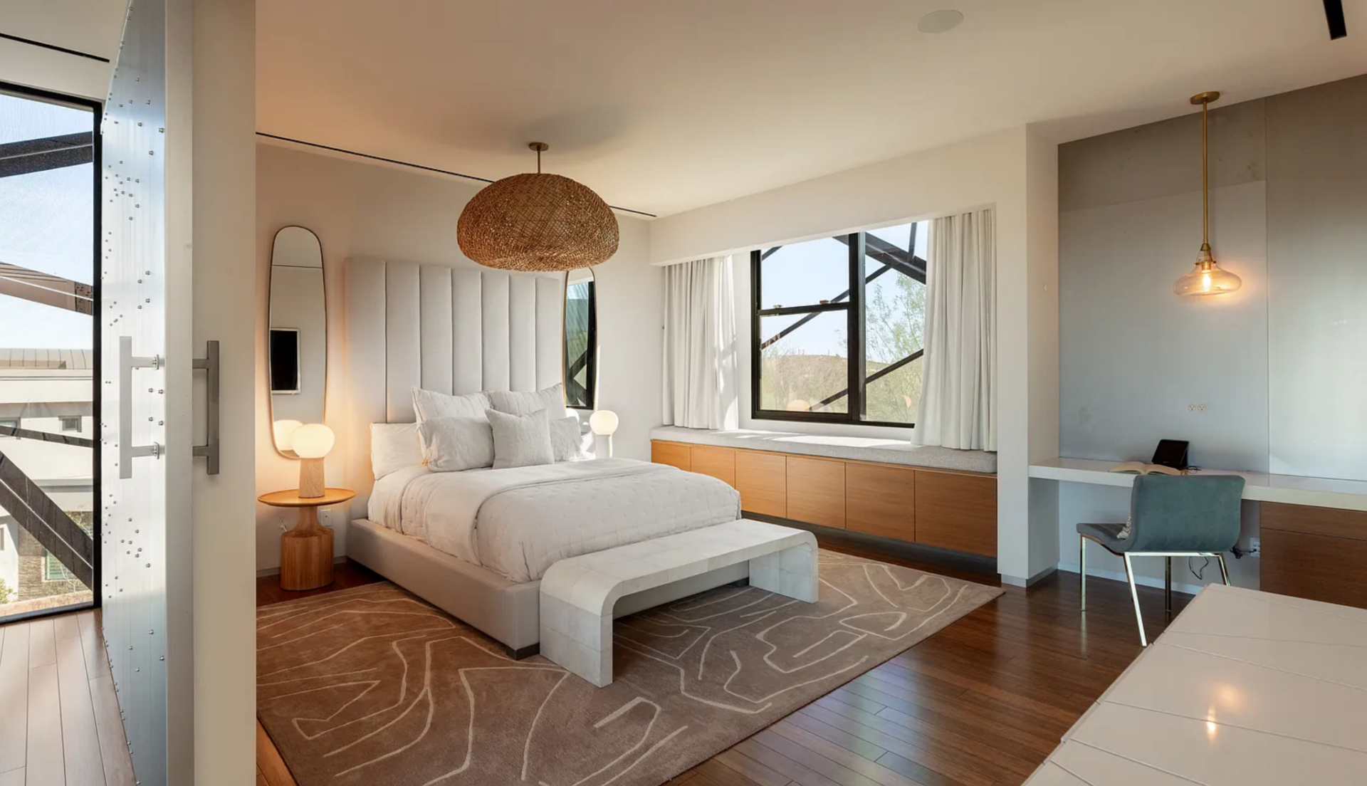 The bedroom centers around a low-profile upholstered bed set against a vertical paneled headboard wall, with a woven pendant overhead adding texture. 