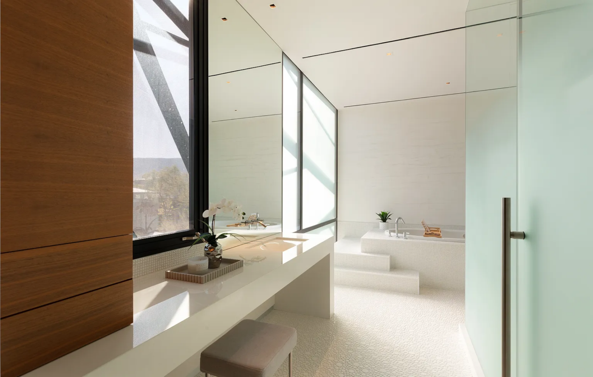 The en-suite bathroom begins with a long vanity that sits beneath a large mirror and window, while the soaking tub is set slightly elevated at the far end, accessed by a short step.