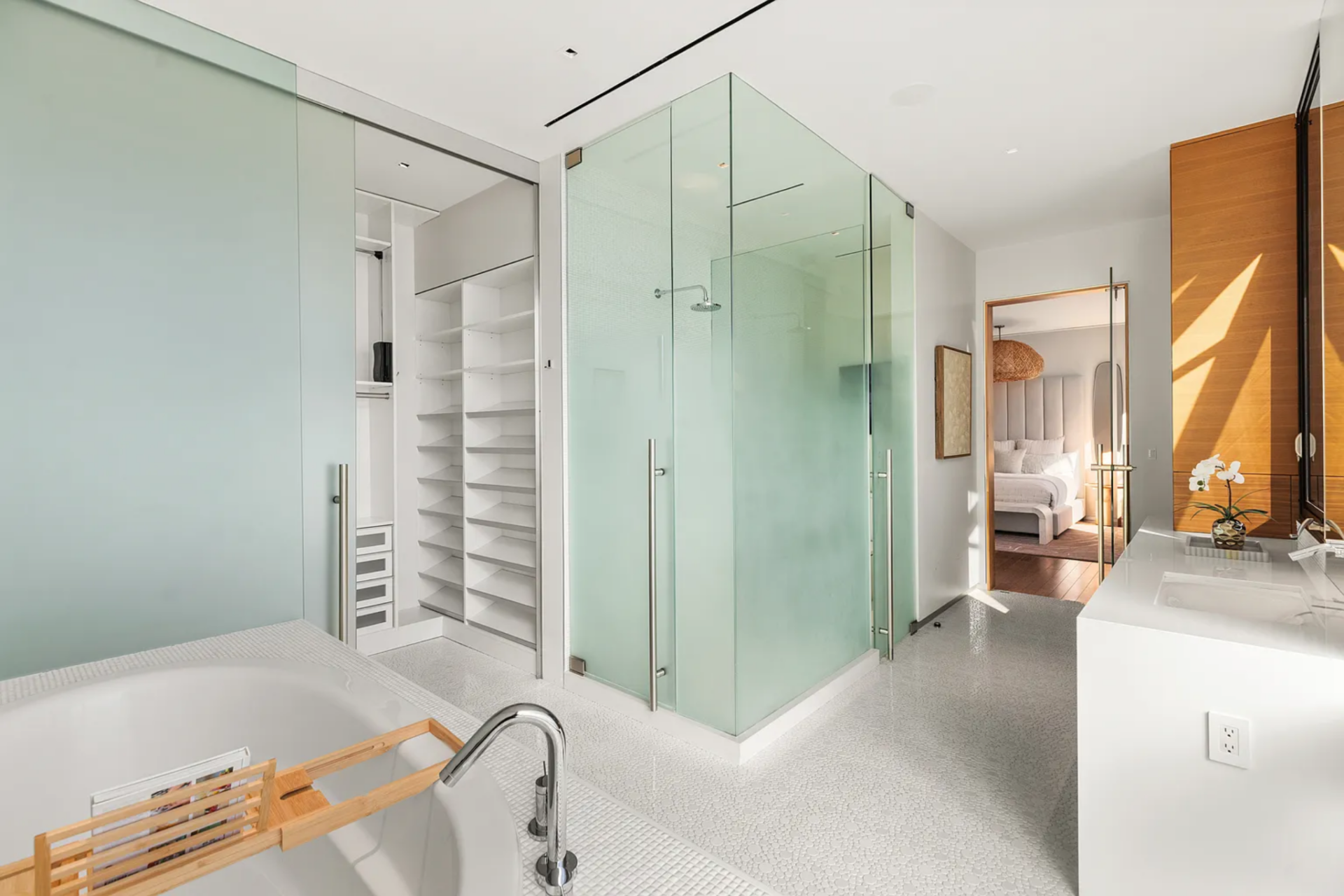 At the center, a glass-enclosed shower sits next to the suite's closet, where open shelving and storage are built directly into the wall.