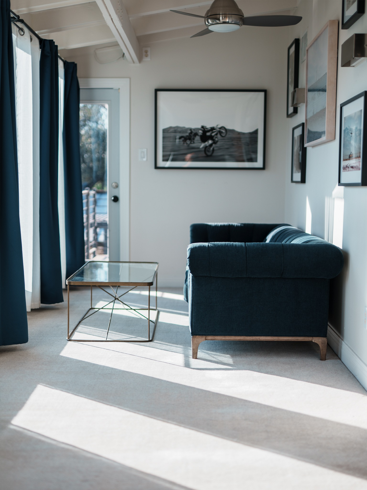 A deep blue velvet sofa, slim metal-and-glass coffee table, and curated gallery wall give the space its own identity, while a nearby glass door provides direct outdoor access.