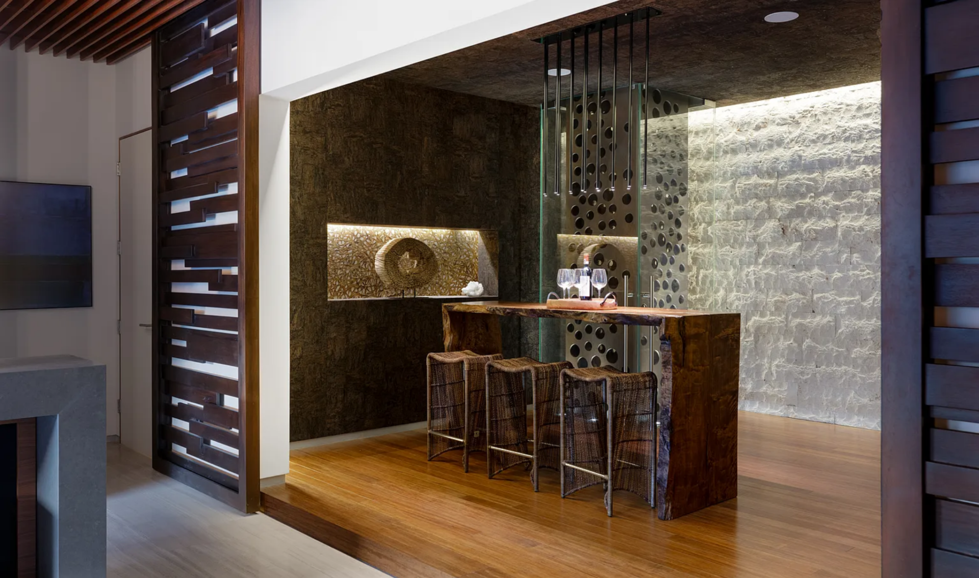 A live-edge wood bar top sits against a backdrop of stone and glass, with integrated display and storage elements built into the wall.